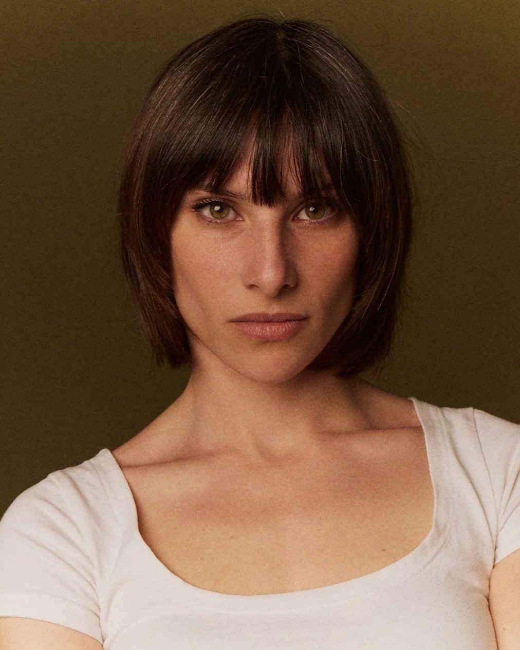Retrato de una mujer con cabello castaño oscuro, ojos claros y expresión seria, usando una camiseta blanca, contra fondo beige.