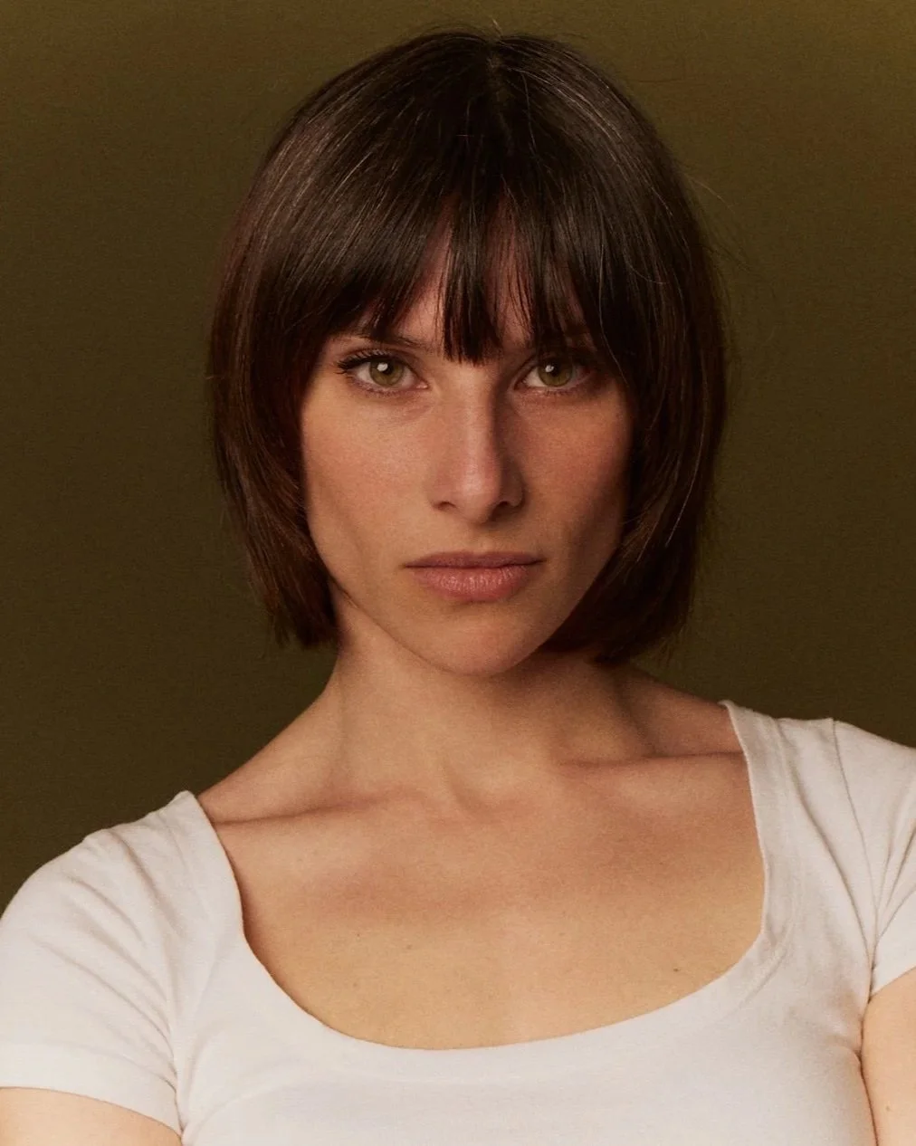 Retrato de una mujer con cabello castaño oscuro y corte de cabello en bob, usando una camiseta blanca con fondo marrón
