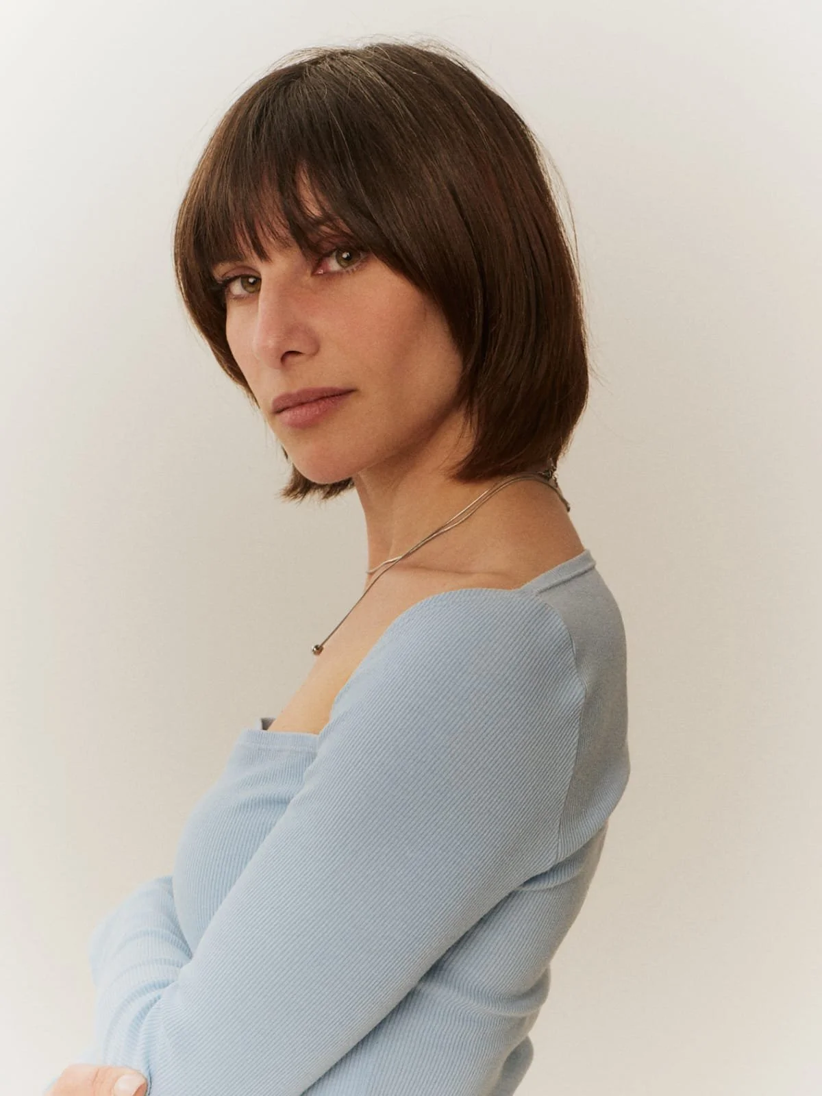 Nina Pomodoro, French-Spanish actress, headshot, Barcelona Retrato de una mujer con cabello castaño oscuro y peinado corto, vestida con una blusa azul y una cadena delgada, posa de perfil frente a un fondo blanco.