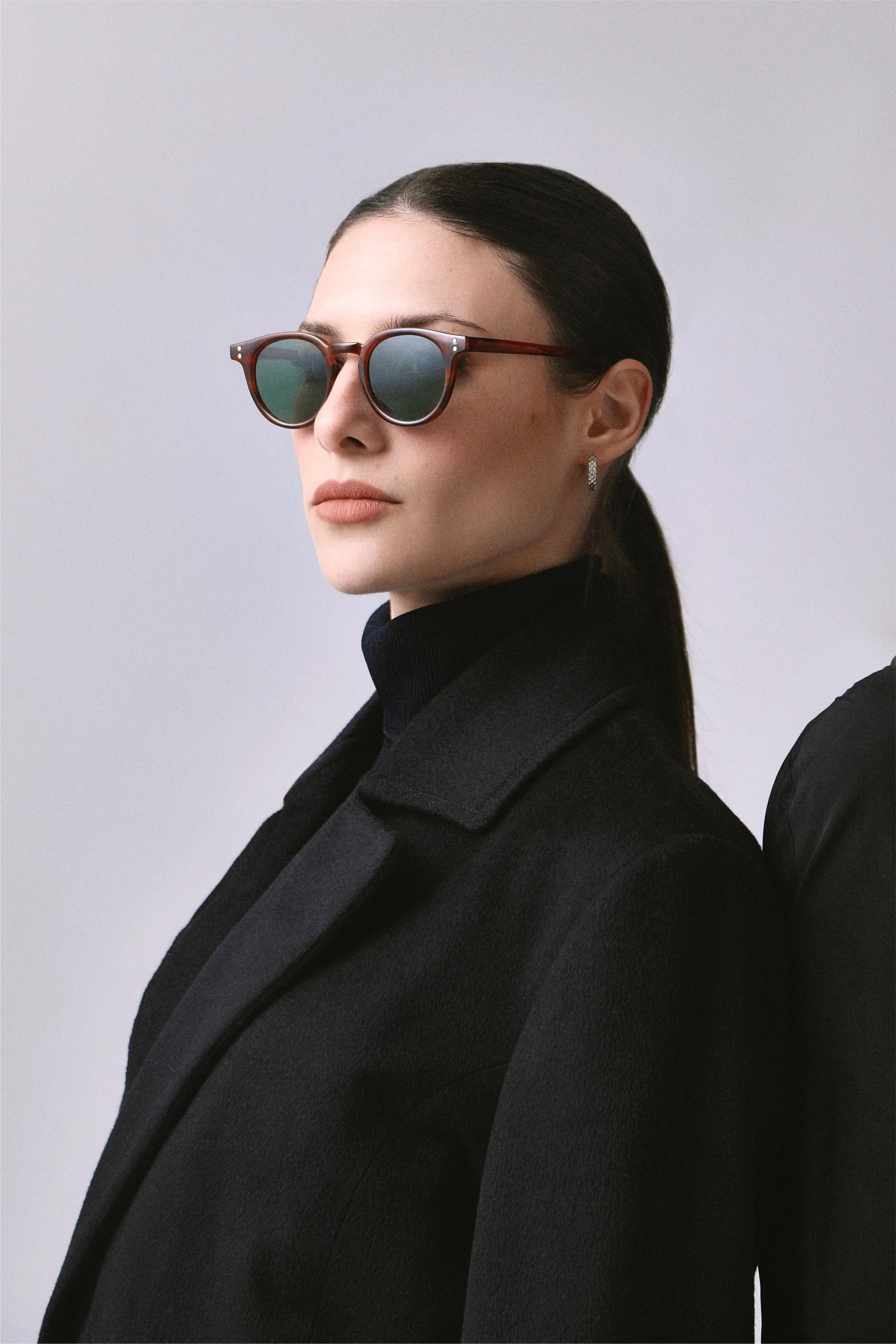 Mujer con cabello recogido, gafas de sol de montura redonda y chaqueta negra de perfil, contra fondo gris.