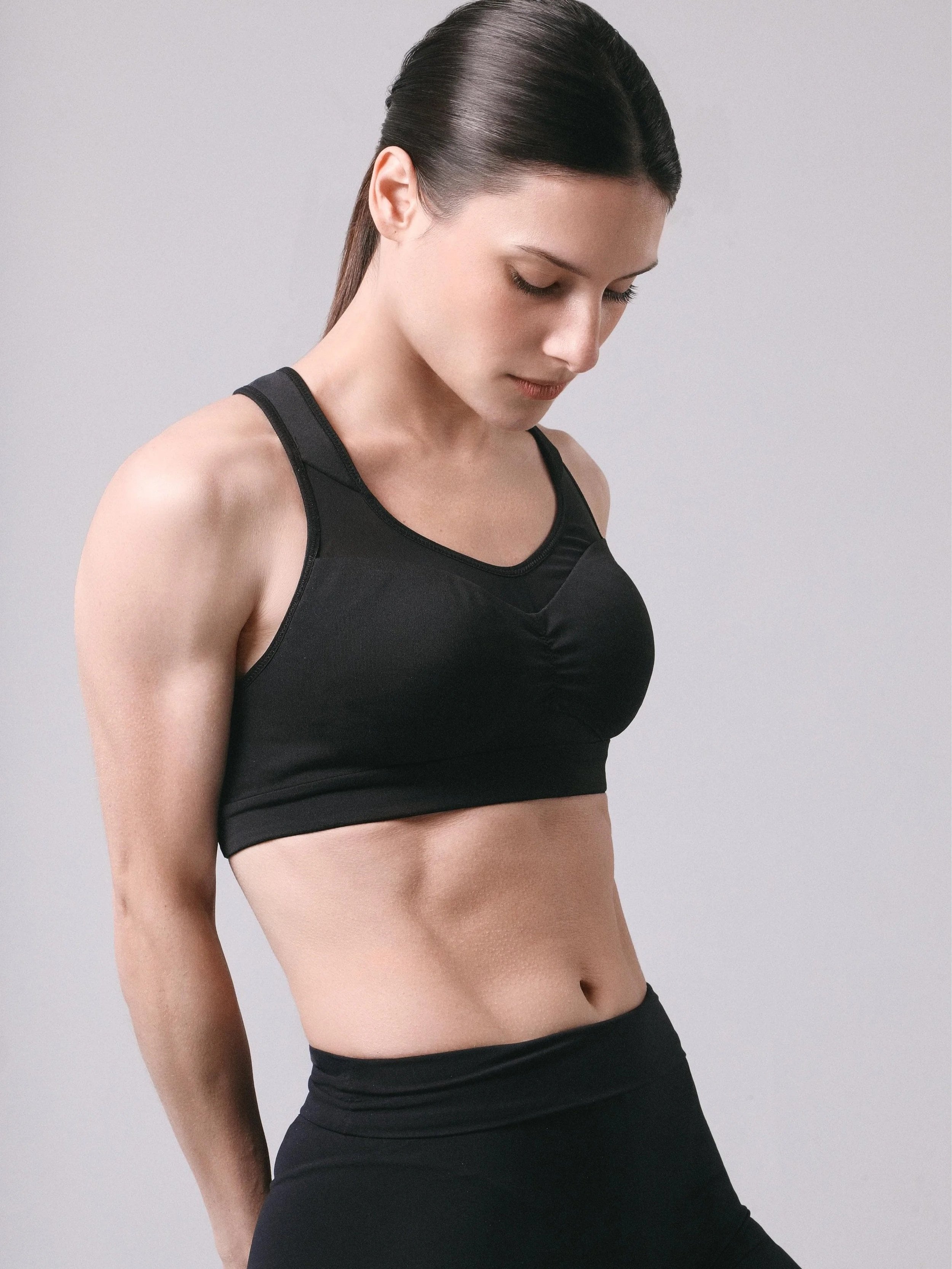Una mujer con blusa deportiva negra y pantalón negro, mirando hacia abajo, con cabello oscuro y liso, fondo gris liso.