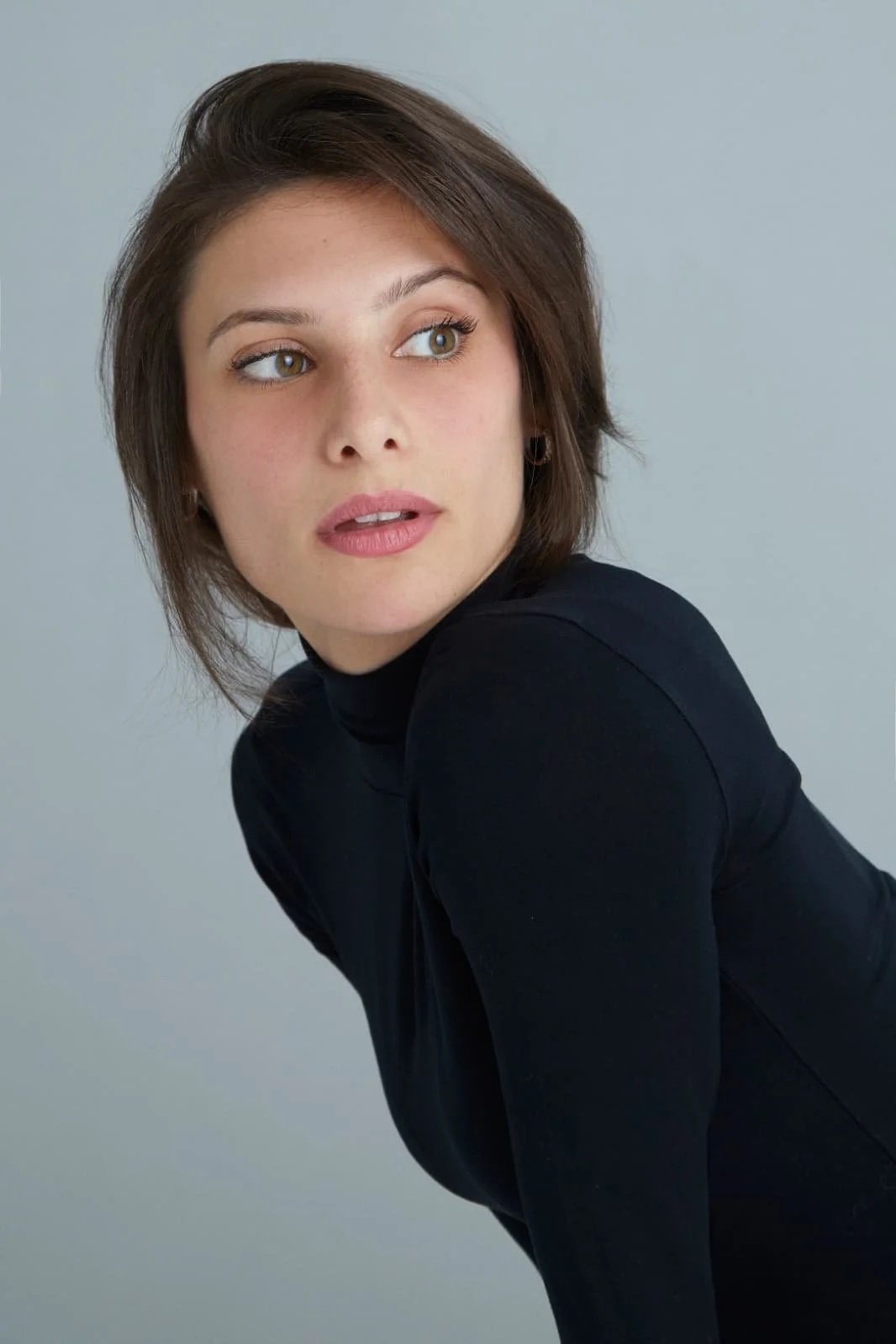 Nina Pomodoro, French-Spanish actress, headshot, Barcelona  Una mujer con cabello castaño oscuro, ojos claros, vestido negro y maquillaje natural, parece estar posando frente a un fondo gris claro.