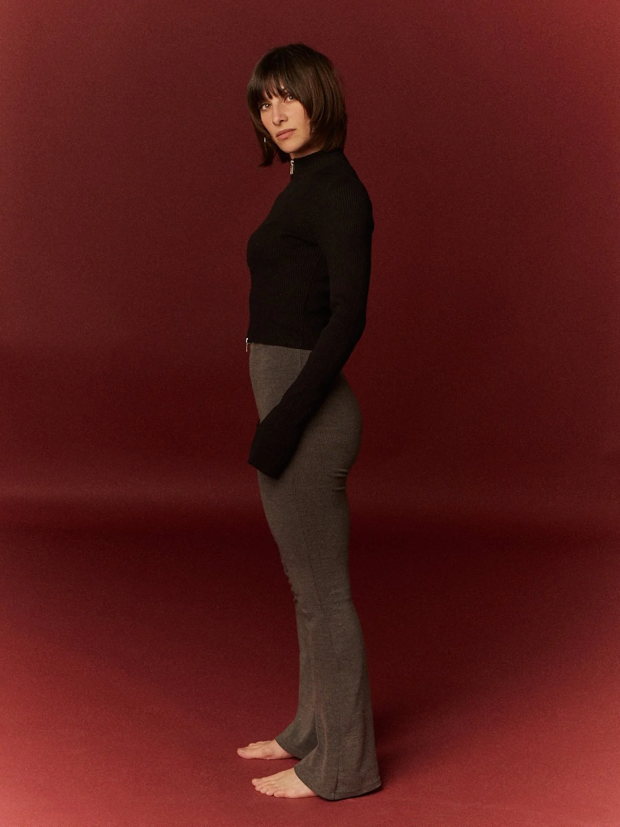 Mujer de cabello castaño corto, con pantalones de tela gris y suéter negro de cuello alto, posando de perfil con fondo rojo oscuro.