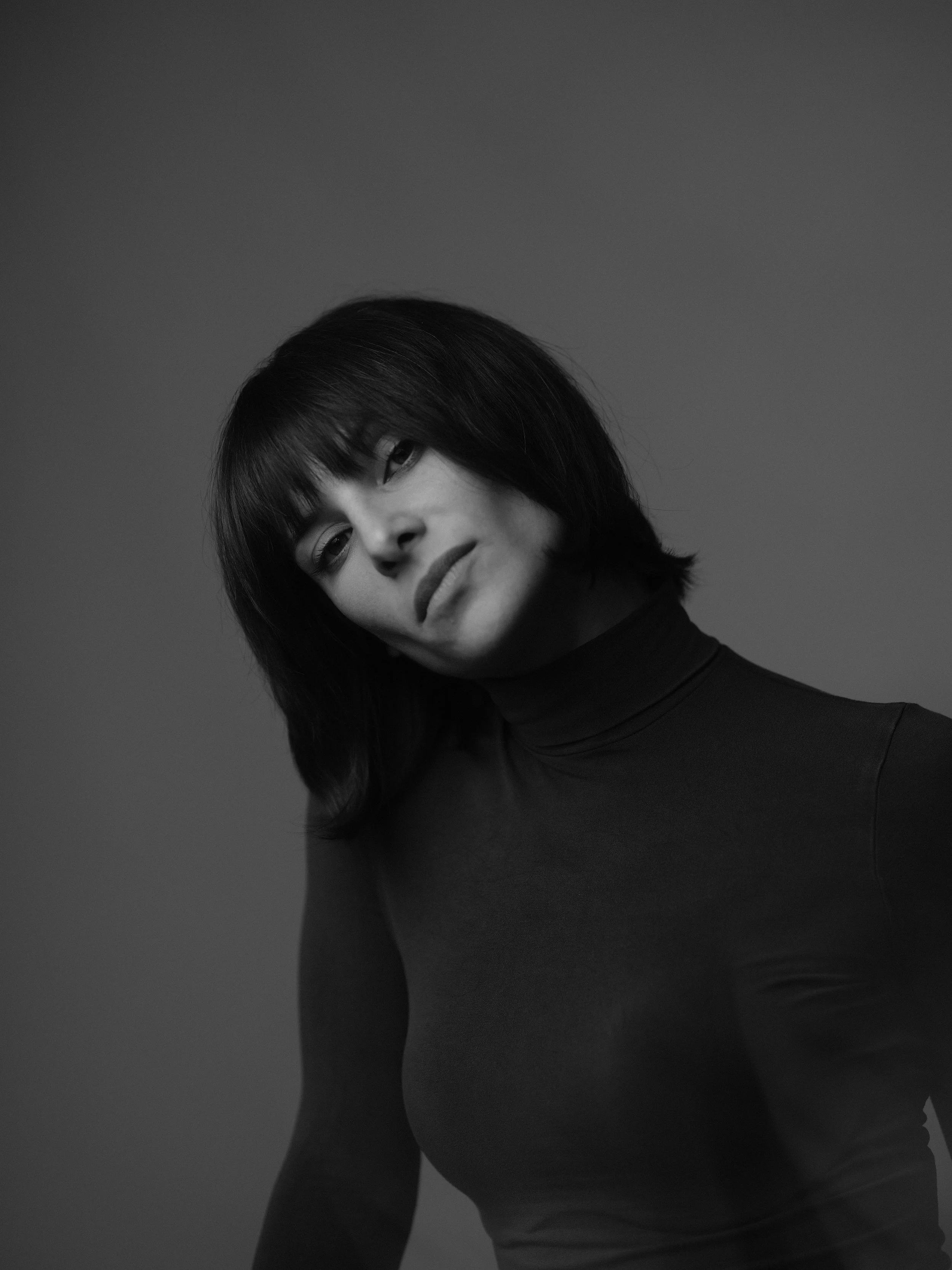 Retrato en blanco y negro de una mujer con cabello rizado y expresión pensativa, vestida con ropa de manga larga, contra un fondo gris.