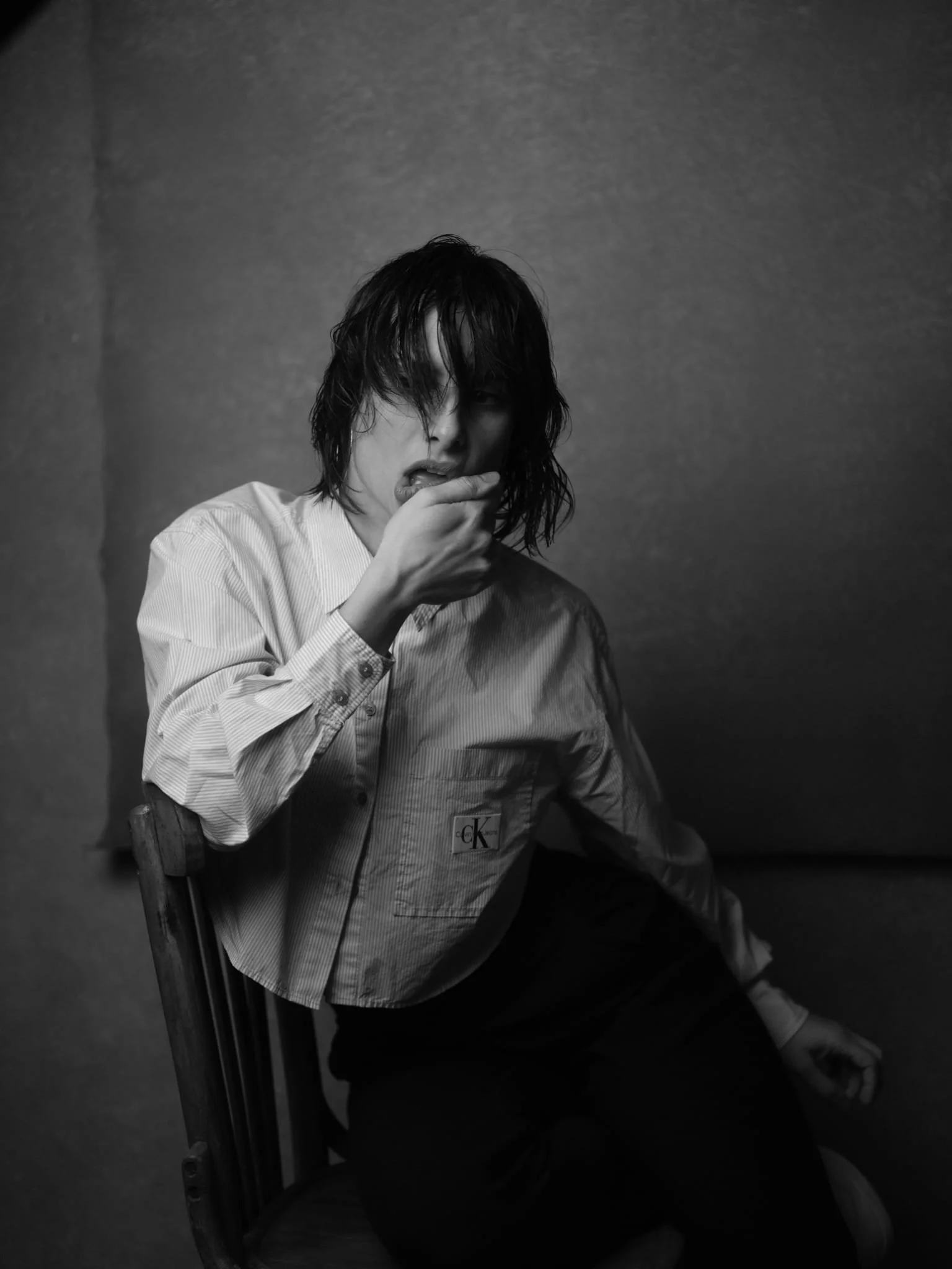 Nina Pomodoro, French-Spanish actress, headshot, Barcelona Mujer con cabello oscuro y mojado, con expresión pensativa, tocándose la boca, vestida con camisa de rayas blancas y negras, sentada en una silla de madera.