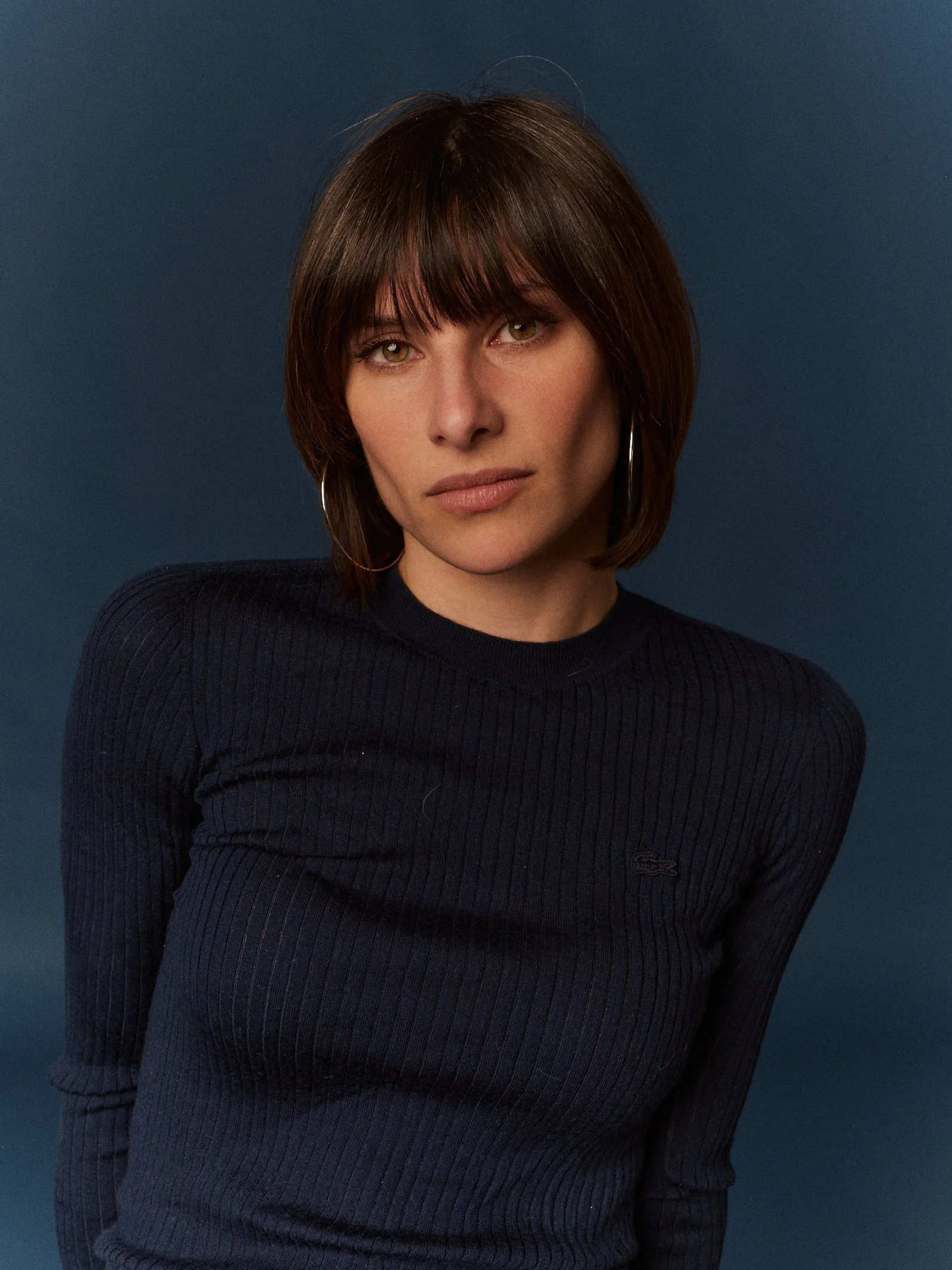 Mujer con cabello castaño oscuro, fleco, abrigo negro con rayas finas, fondo azul oscuro.
