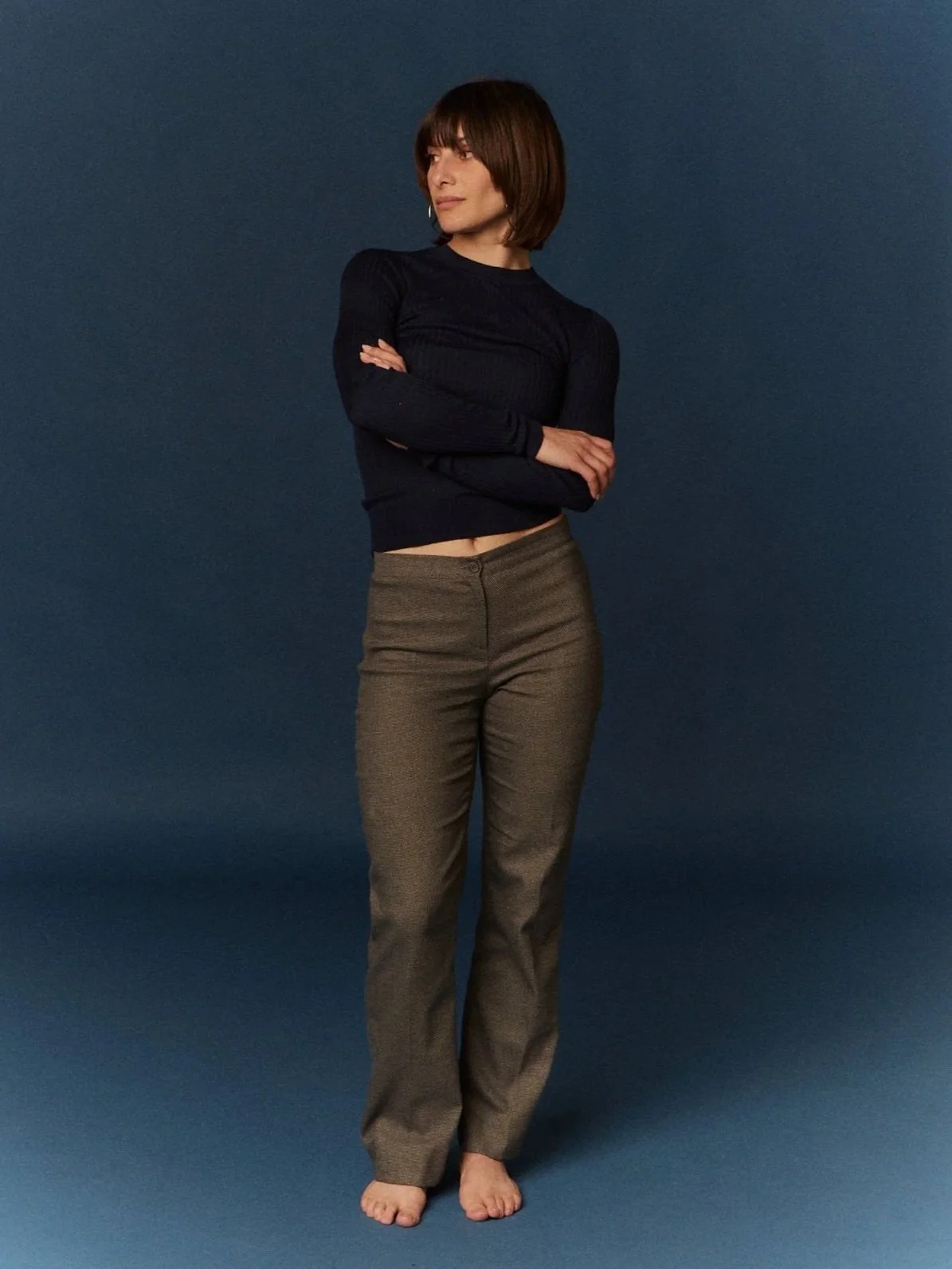 Mujer con cabello castaño oscuro y peinado bob, vestida con blusa negra de manga larga y pantalones beige, de pie con brazos cruzados frente a un fondo azul oscuro.