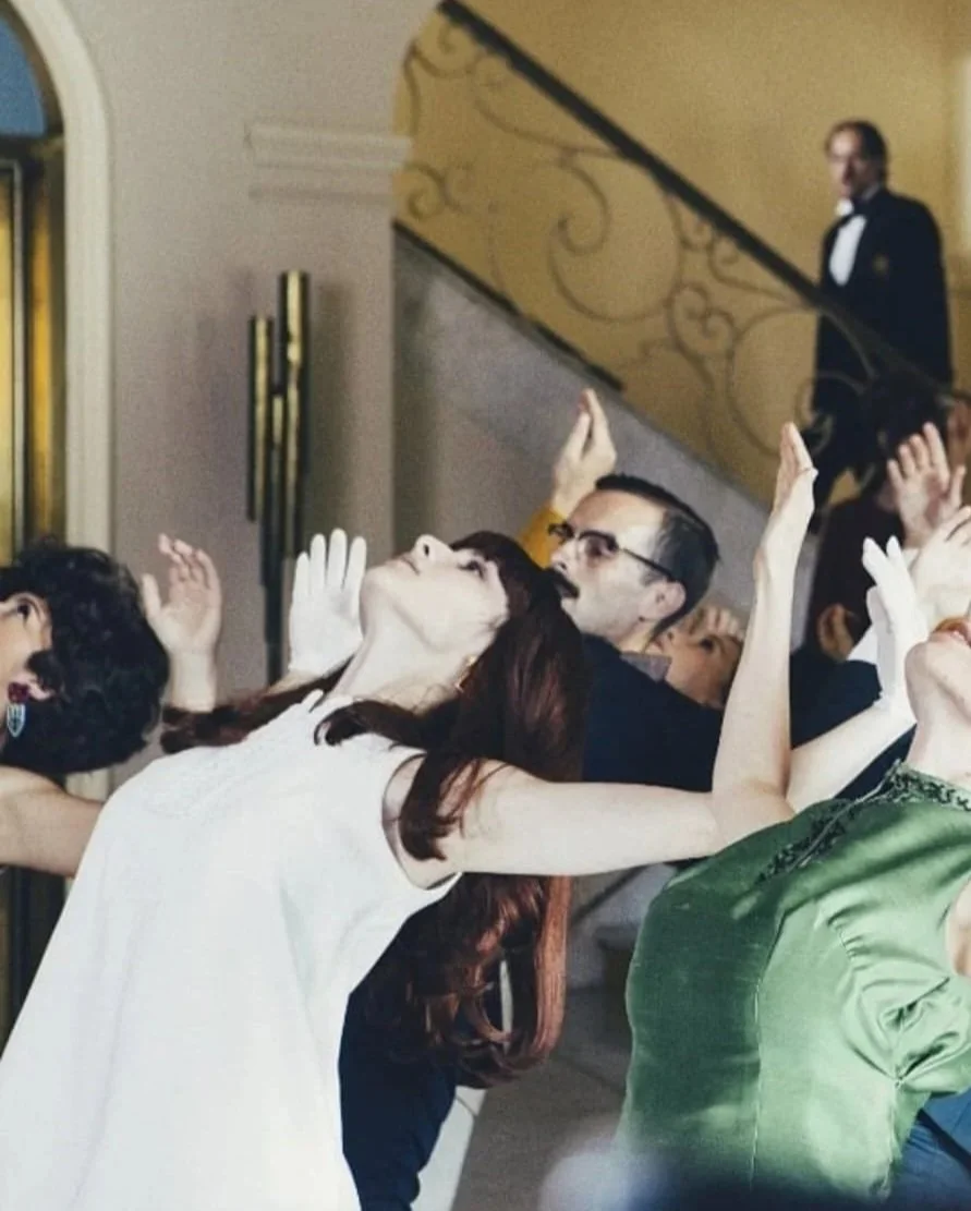 Una escena en un evento social con varias personas bailando y levantando las manos, en un interior elegante con una escalera dorada al fondo y un botones o guardia en uniforme en la parte superior de la escalera.