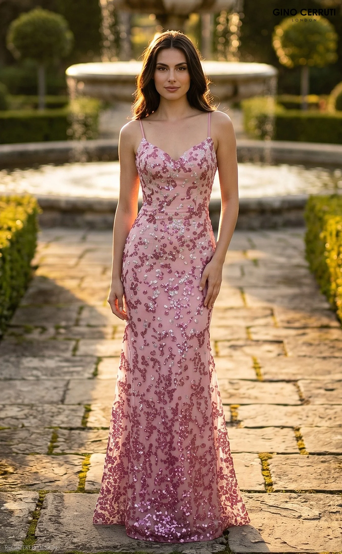 A woman with long dark hair in a pink sequined gown standing on a stone pathway near a fountain in a garden at sunset.