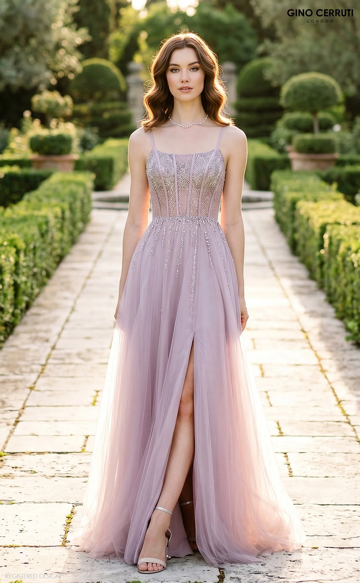 A woman in a long, lavender evening gown standing on a stone pathway in a garden with trimmed bushes and trees. The gown is embellished with silver details and has a slit revealing her leg. She is wearing high heels, a pearl necklace, and has loose w