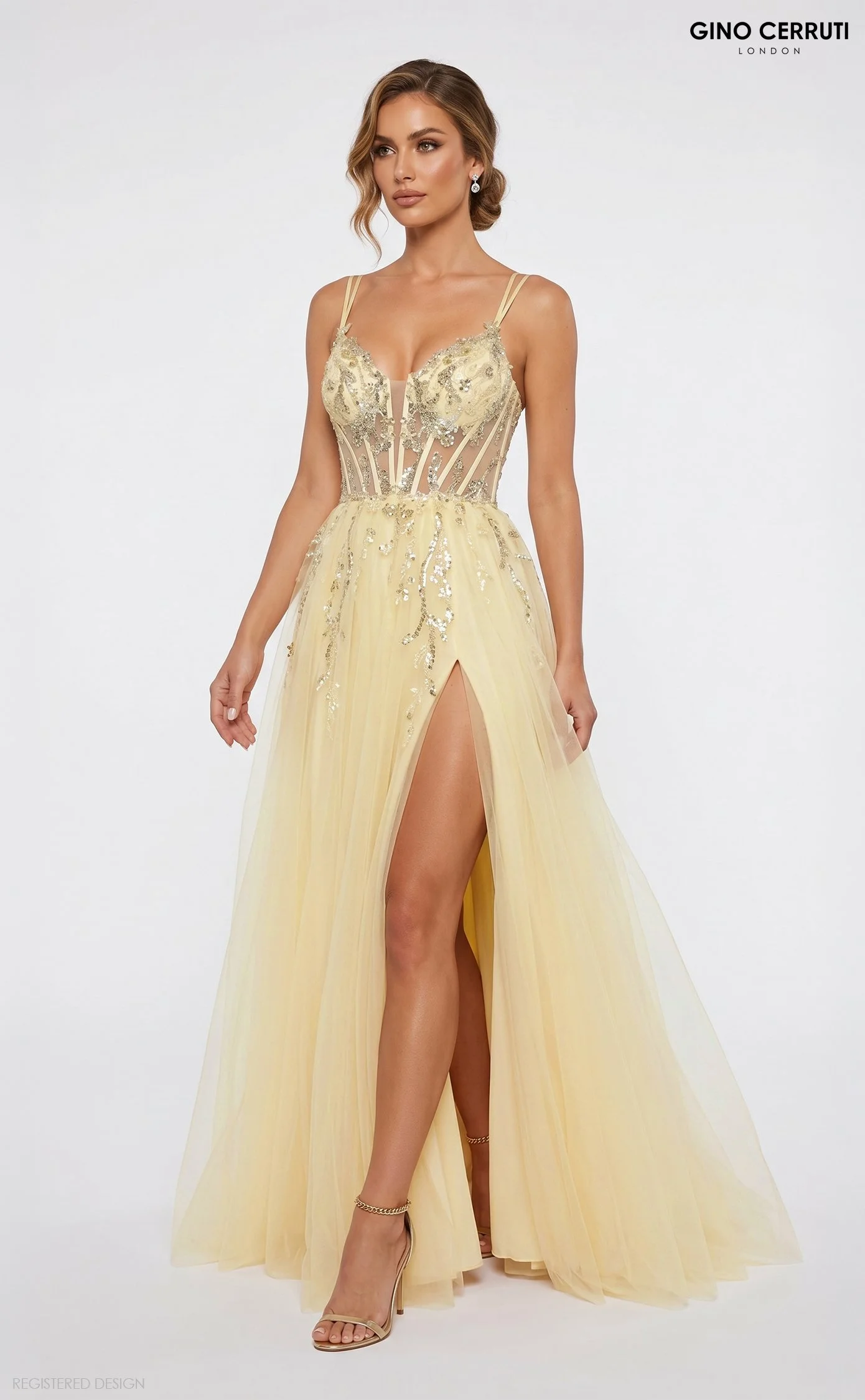 A woman wearing a yellow evening gown with a high slit, intricate gold embroidery, and a sheer bodice, standing against a white background.