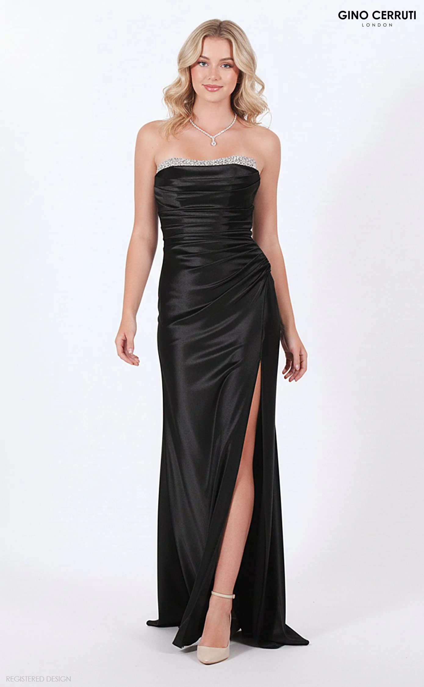 A woman in a strapless, black satin gown with a thigh-high slit, wearing beige heels, a diamond necklace, and posing against a plain white background. The top of the image has the text 'Gino Cerruti London'.