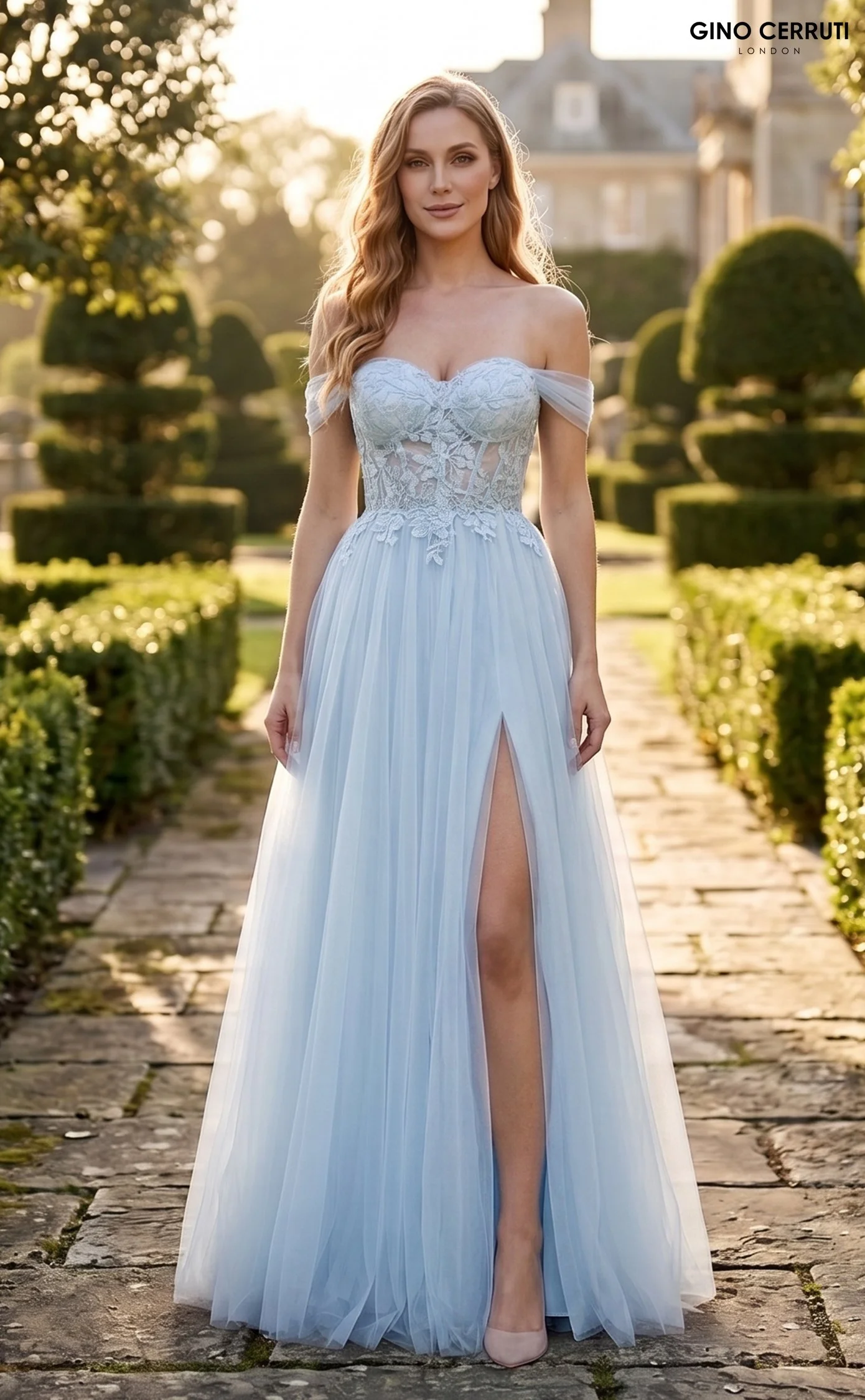 A woman wearing a light blue, off-shoulder, lace wedding dress with a slit on the front, standing on a cobblestone pathway in a garden with trimmed bushes and trees, sunset lighting.