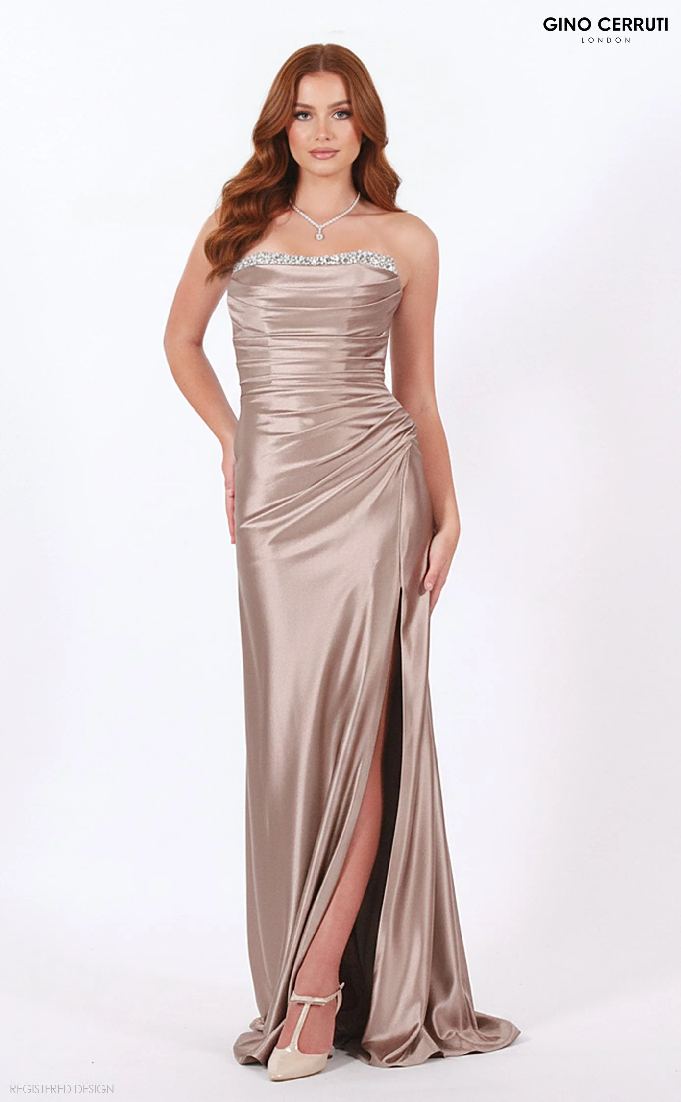 A woman with long red hair wearing a strapless, champagne-colored satin gown with a slit, standing against a plain white background, accessorized with a diamond necklace and high heels.