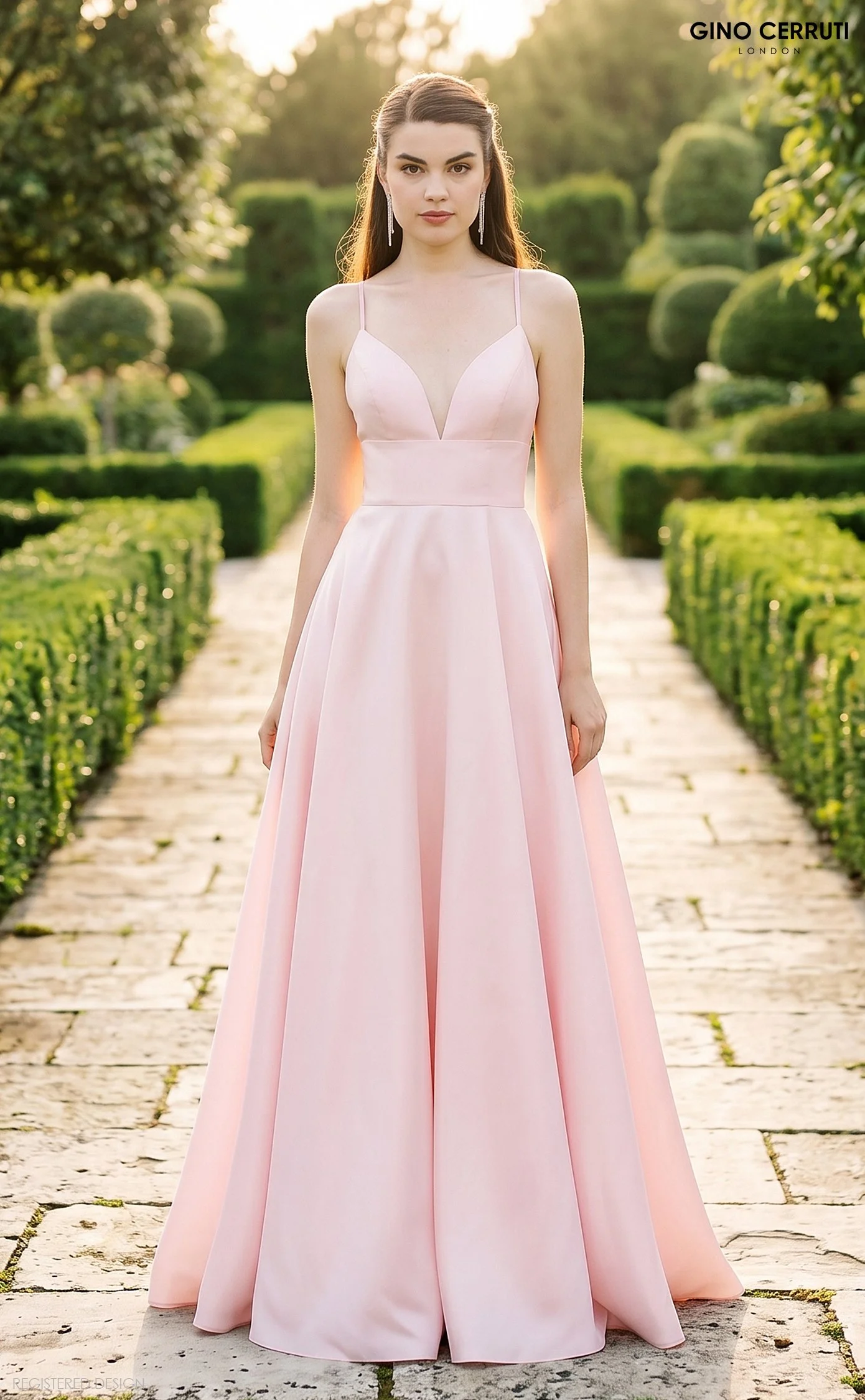A woman in a long, light pink evening gown standing on a stone pathway in a garden, with trees and neatly trimmed hedges in the background during sunset.