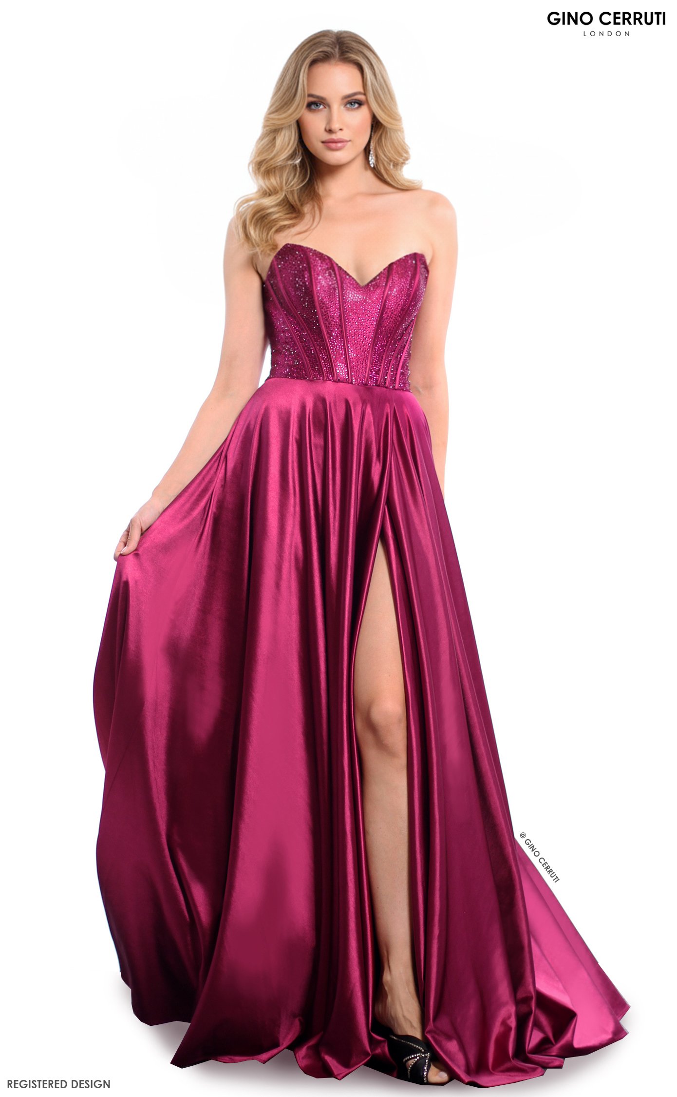 A woman with blonde hair wearing a strapless, deep pink gown with a slit revealing black high-heeled shoes. The dress has a fitted, embellished bodice and a flowing, shiny skirt. The image is a fashion photo with a plain white background.