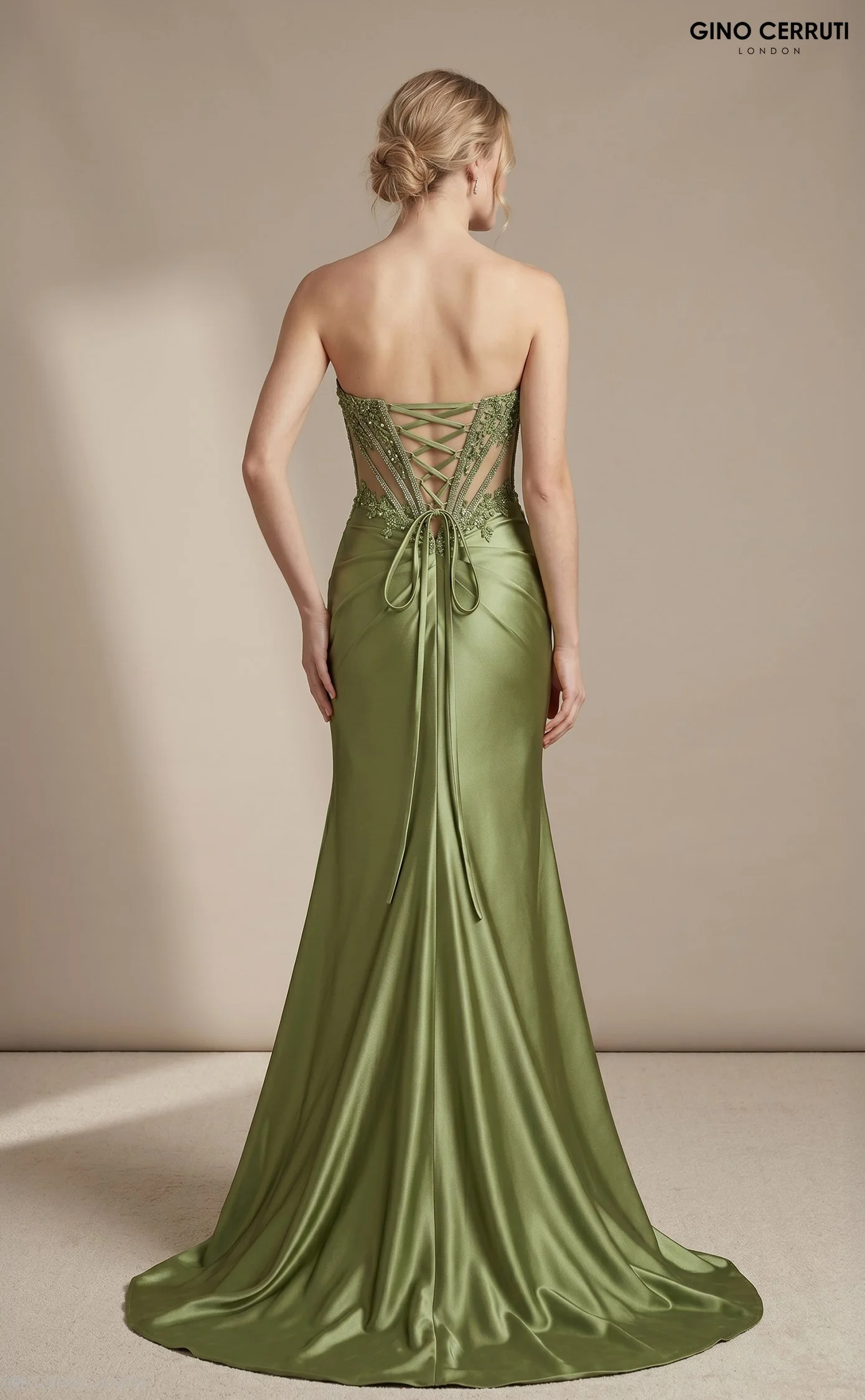 Back view of a woman wearing a green satin gown with intricate beaded embroidery and a lace-up back detail. The gown has a fitted bodice and a flowing skirt. The woman has blonde hair styled in an elegant bun and is standing against a neutral backgro