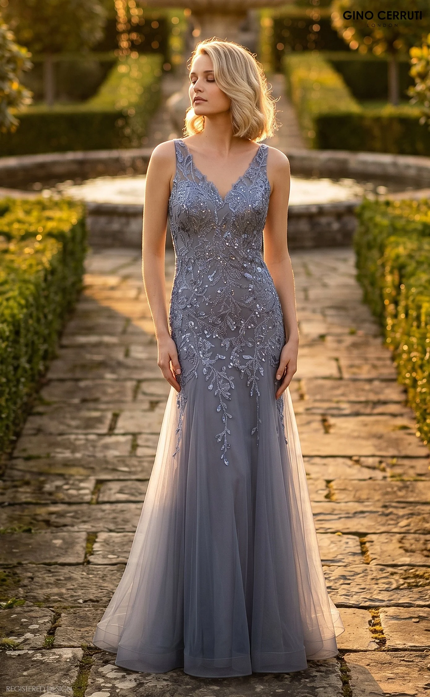 A woman in a light gray lace and beaded evening gown standing on a cobblestone pathway in a garden during sunset, with her eyes closed and a serene expression.