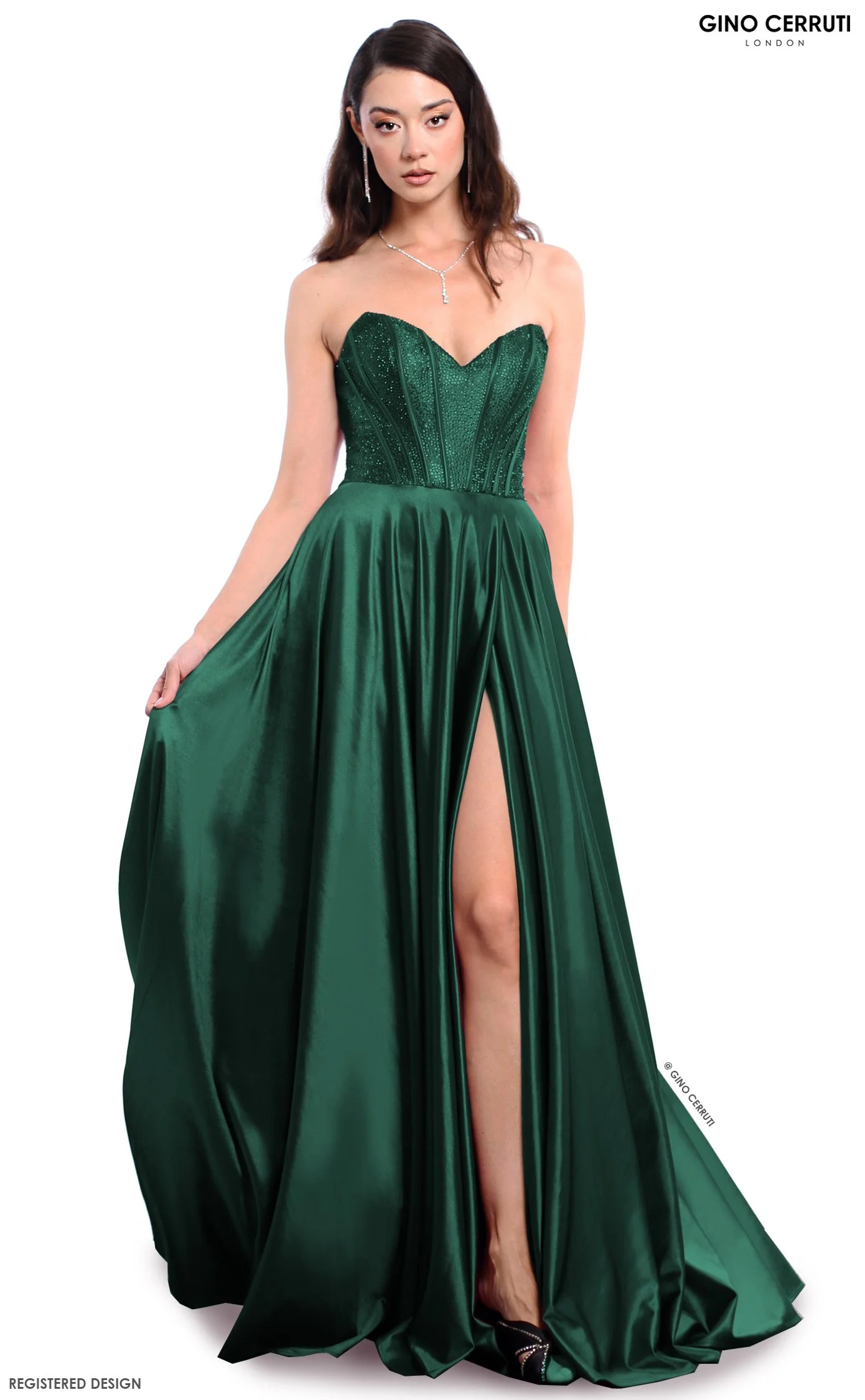 A woman in an emerald green strapless evening gown with a high slit, wearing jewelry including earrings and a necklace, and standing against a white background. The dress is fitted at the top with a sweetheart neckline and flowing at the bottom. The 