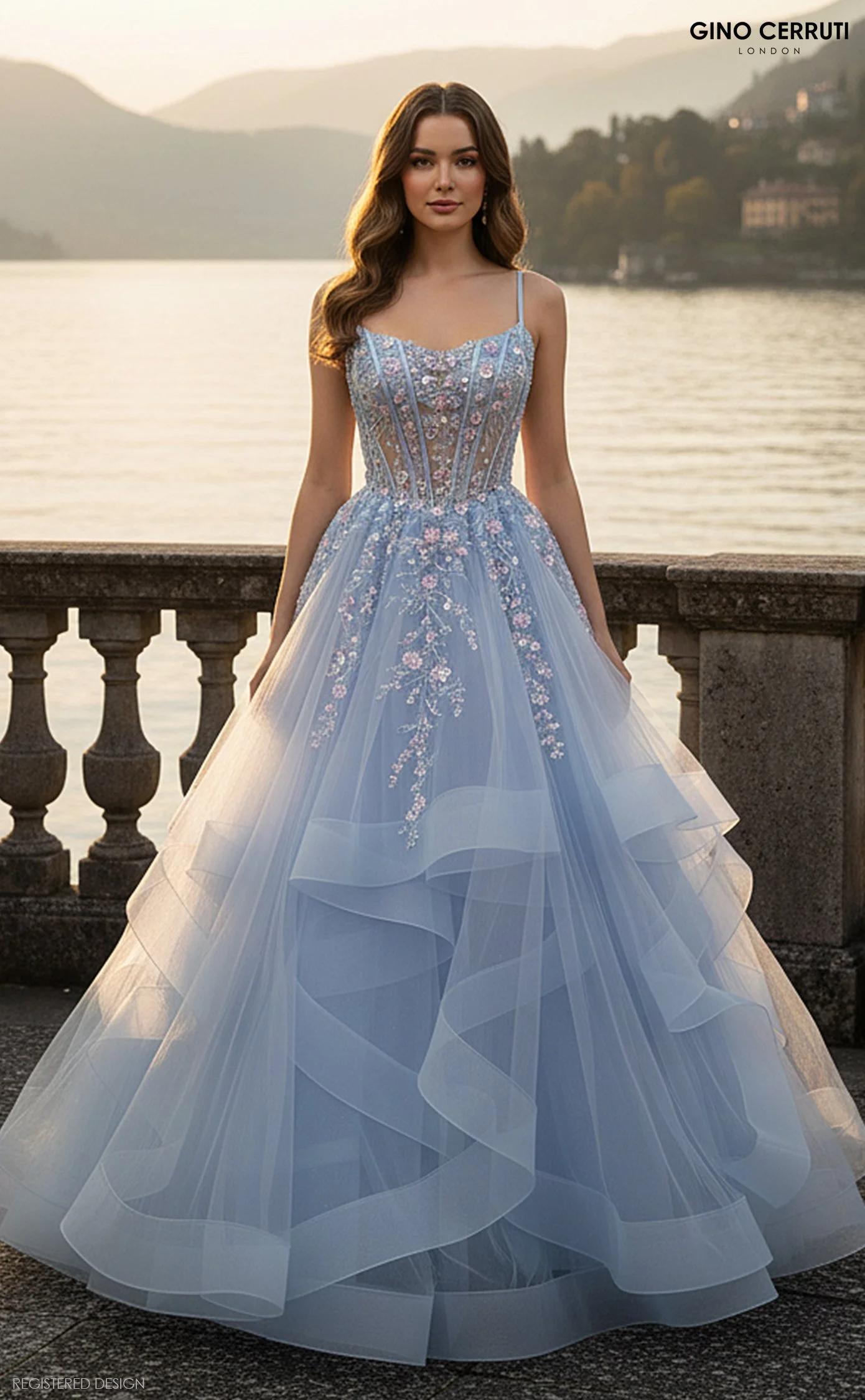 A young woman in a light blue wedding gown with floral embroidery stands by a stone balustrade overlooking a lake at sunset, with mountains in the background.