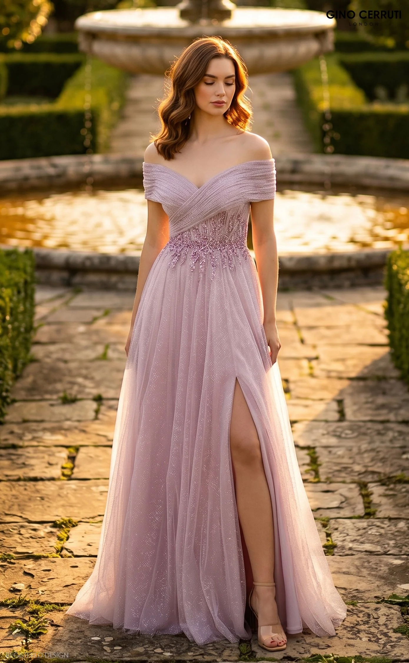 A woman in a light pink off-shoulder gown standing outdoors on a stone pathway next to a fountain, during sunset.