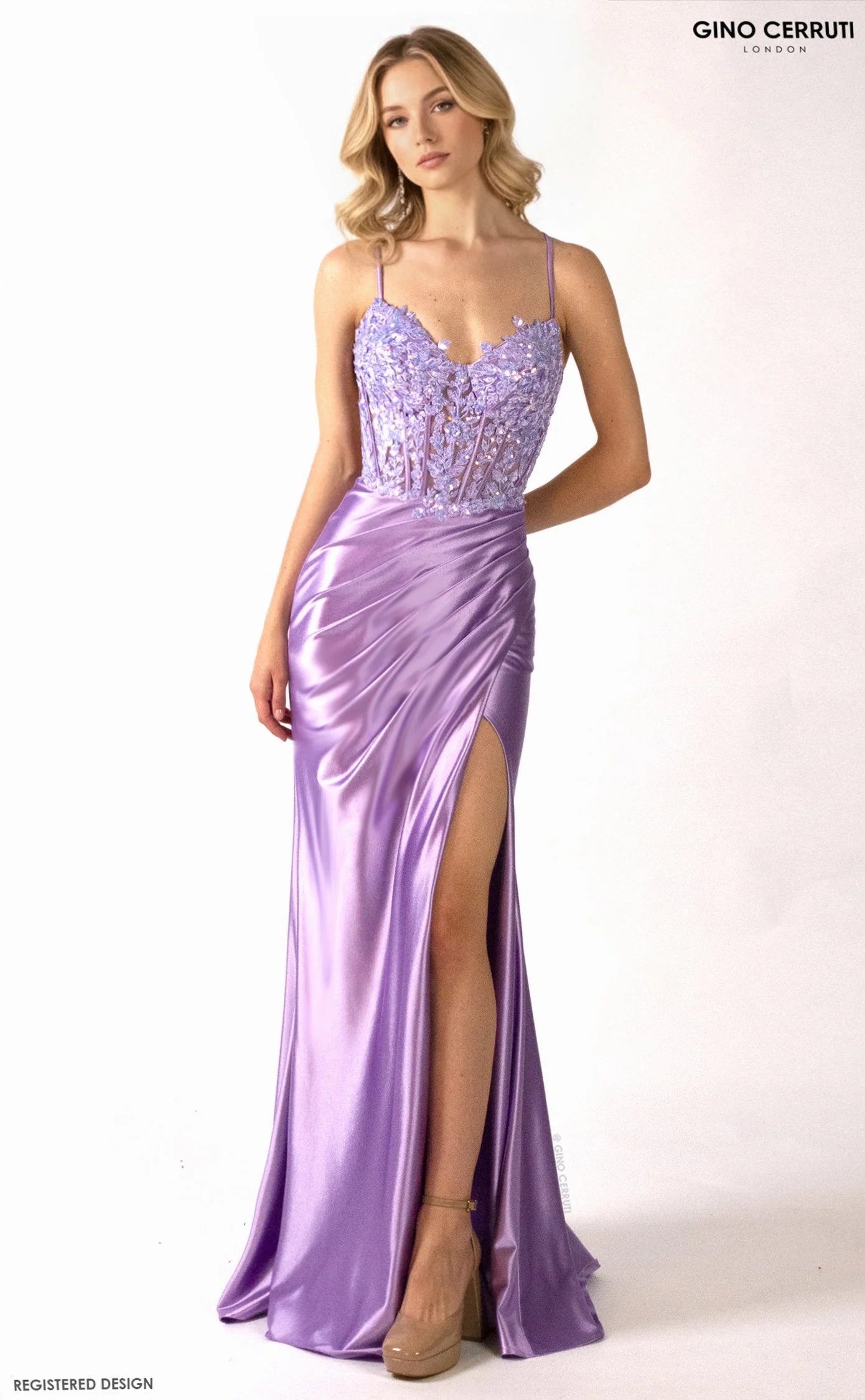 A young woman with blonde wavy hair modeling a lavender satin evening gown with a thigh-high slit and a fitted, embroidered bodice. She is wearing beige platform heels and poses against a white background.