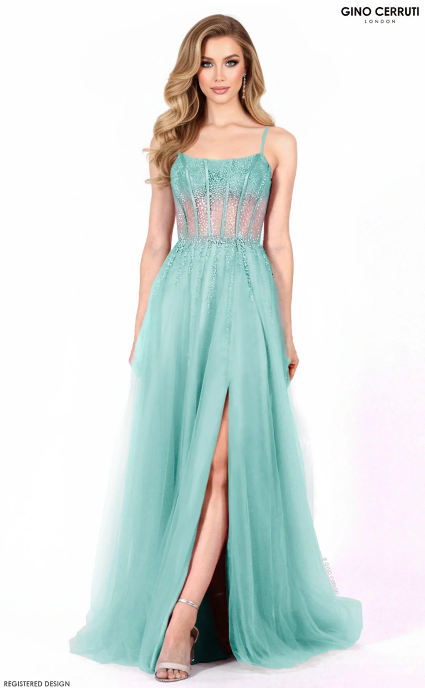 A woman with wavy blonde hair and makeup wearing a turquoise evening gown with spaghetti straps, sheer and embellished bodice, and a high slit, standing against a plain white background.