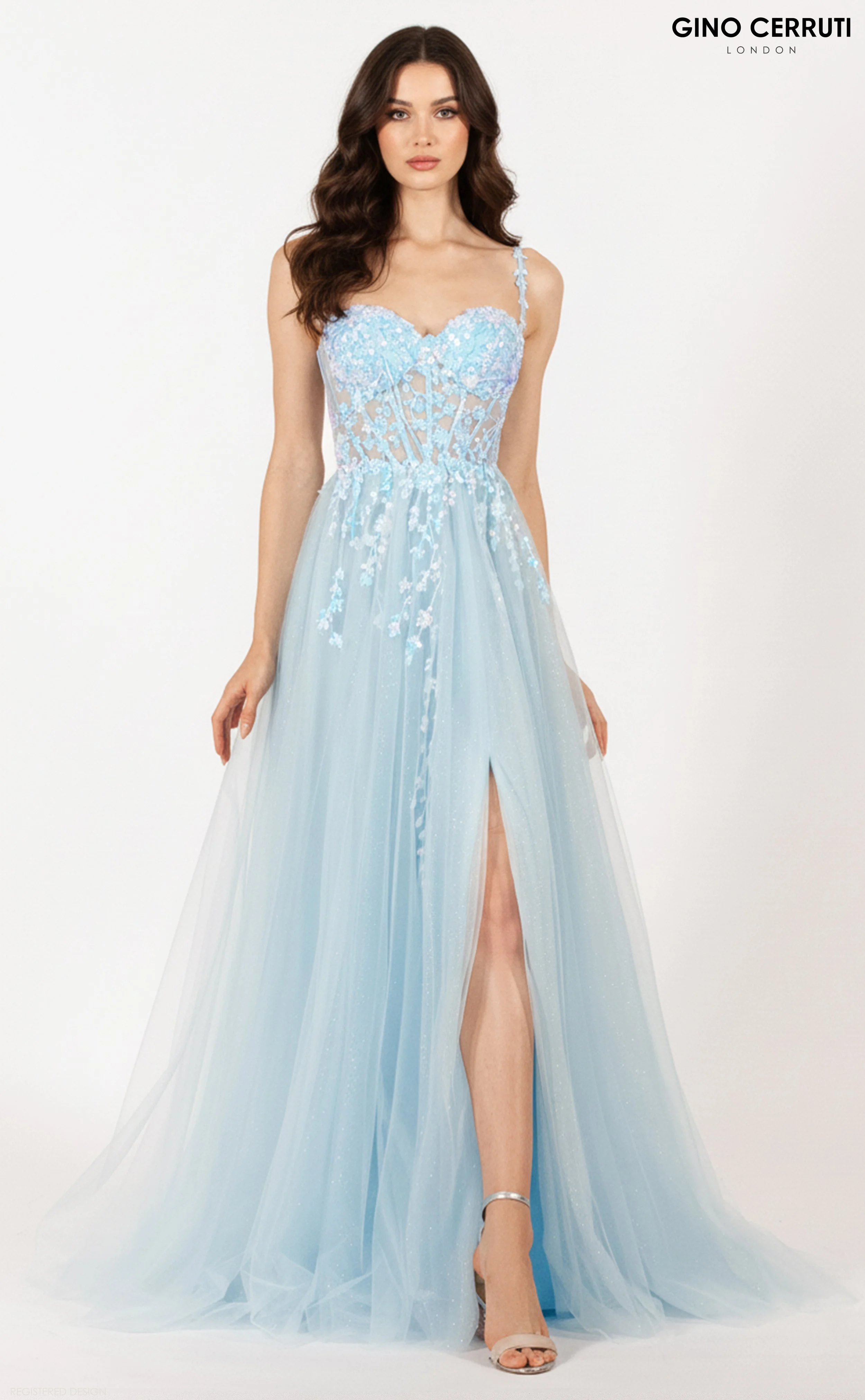 A woman in a light blue, floral embroidery gown with a thigh-high slit, standing against a plain white background. The dress has thin straps and a fitted bodice, with her long dark hair styled in loose waves. She is wearing silver open-toe high heels