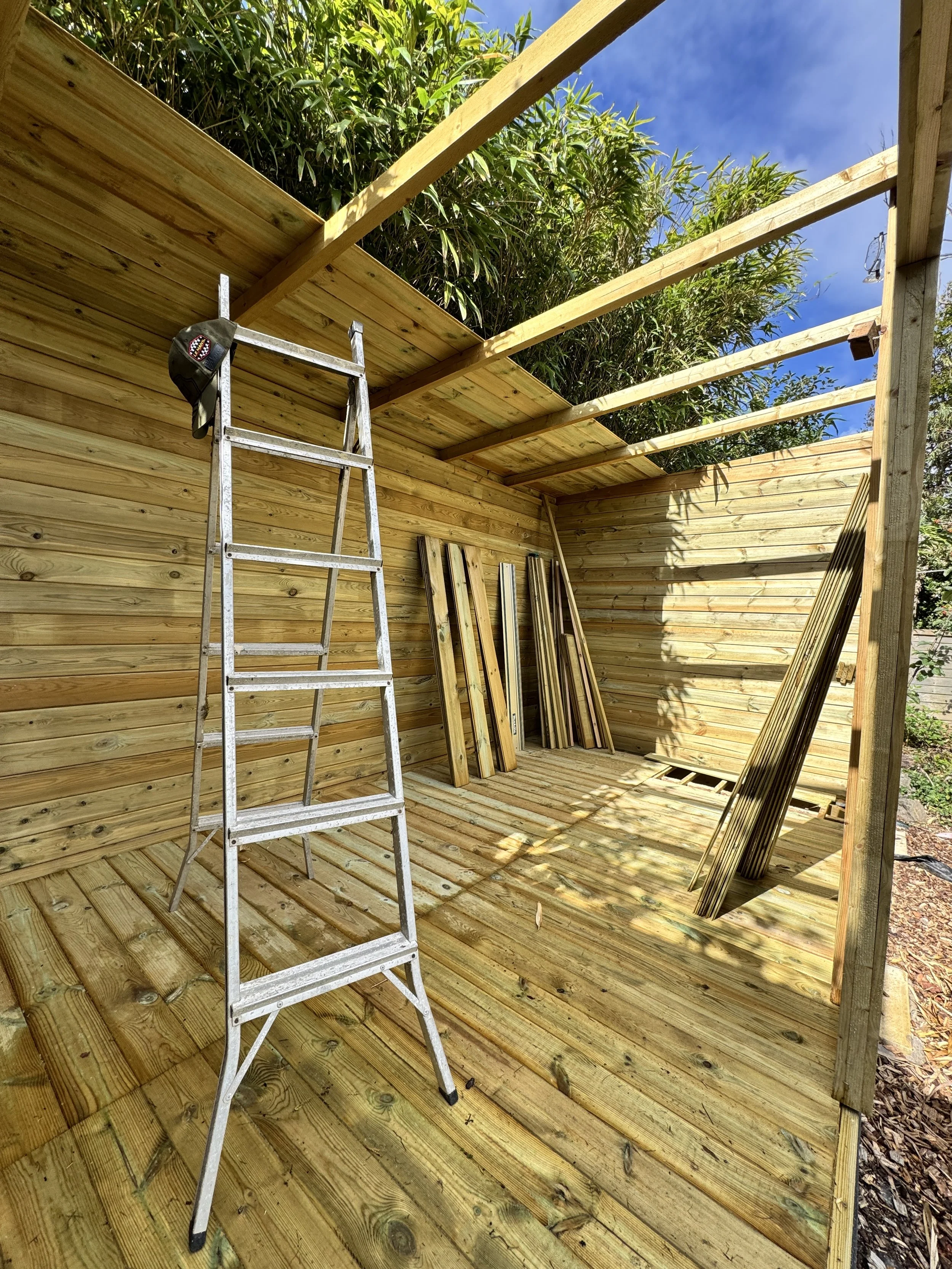 This image shows a partially constructed outdoor wooden shed or similar structure with a metal step ladder inside, leaning against one wall. Several wooden planks are stacked against the wall and leaning on the floor, and there are green trees and a blue sky in the background.