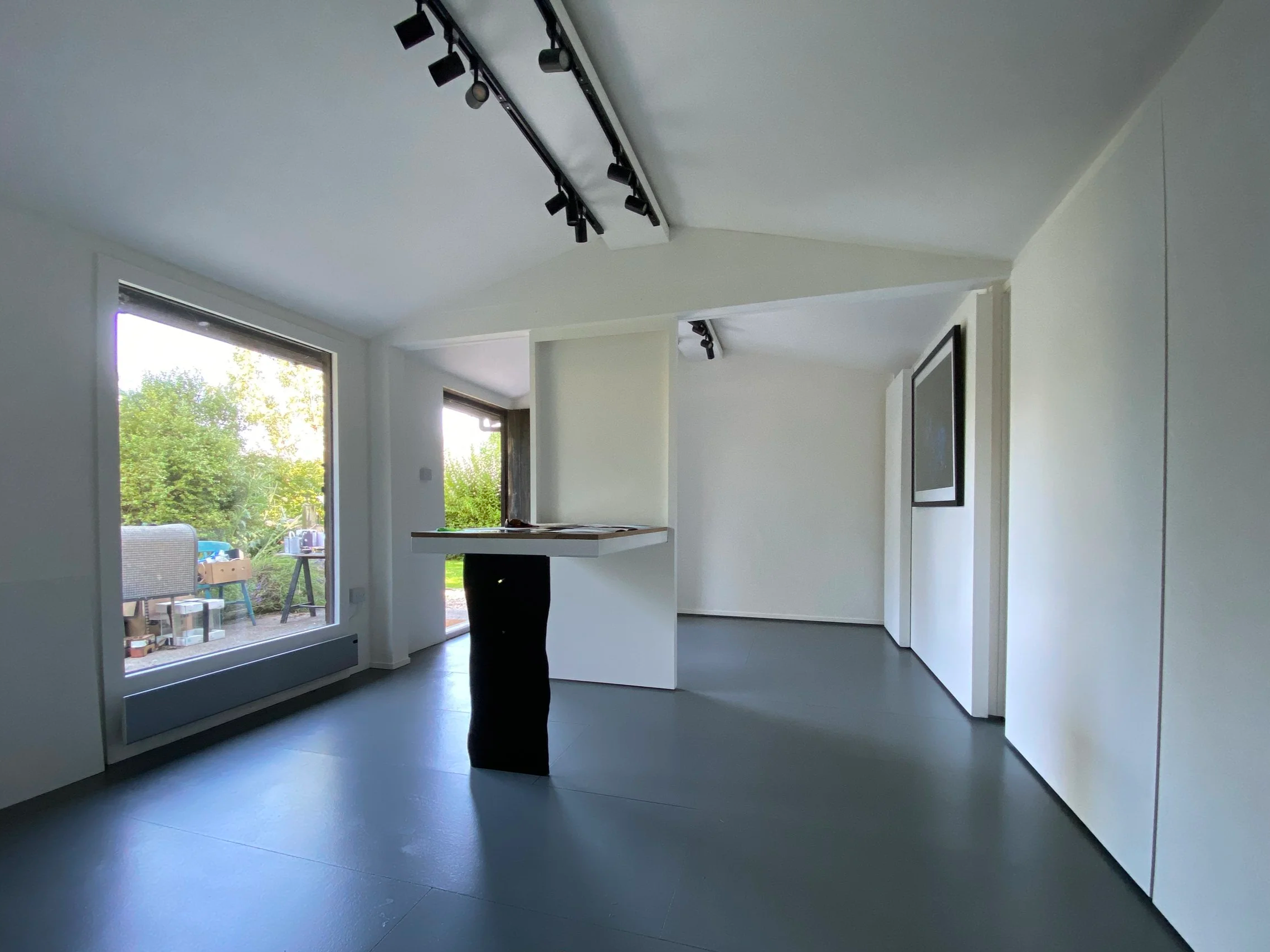 Interior of a modern home with large open space, dark flooring, white walls, and a ceiling track lighting system. There are large windows bringing in natural light, with an outdoor area visible through the glass, including outdoor furniture and greenery.