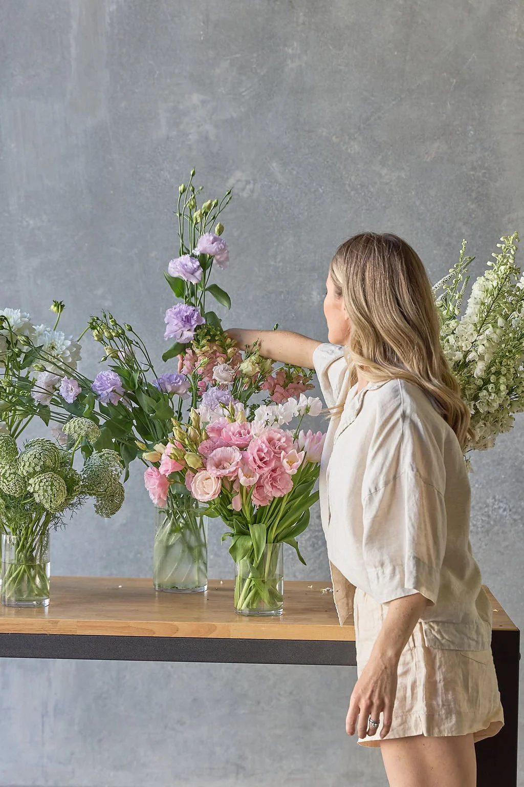 Woman arranging pastel-colored flowers in glass vases on a wooden table against a gray wall.