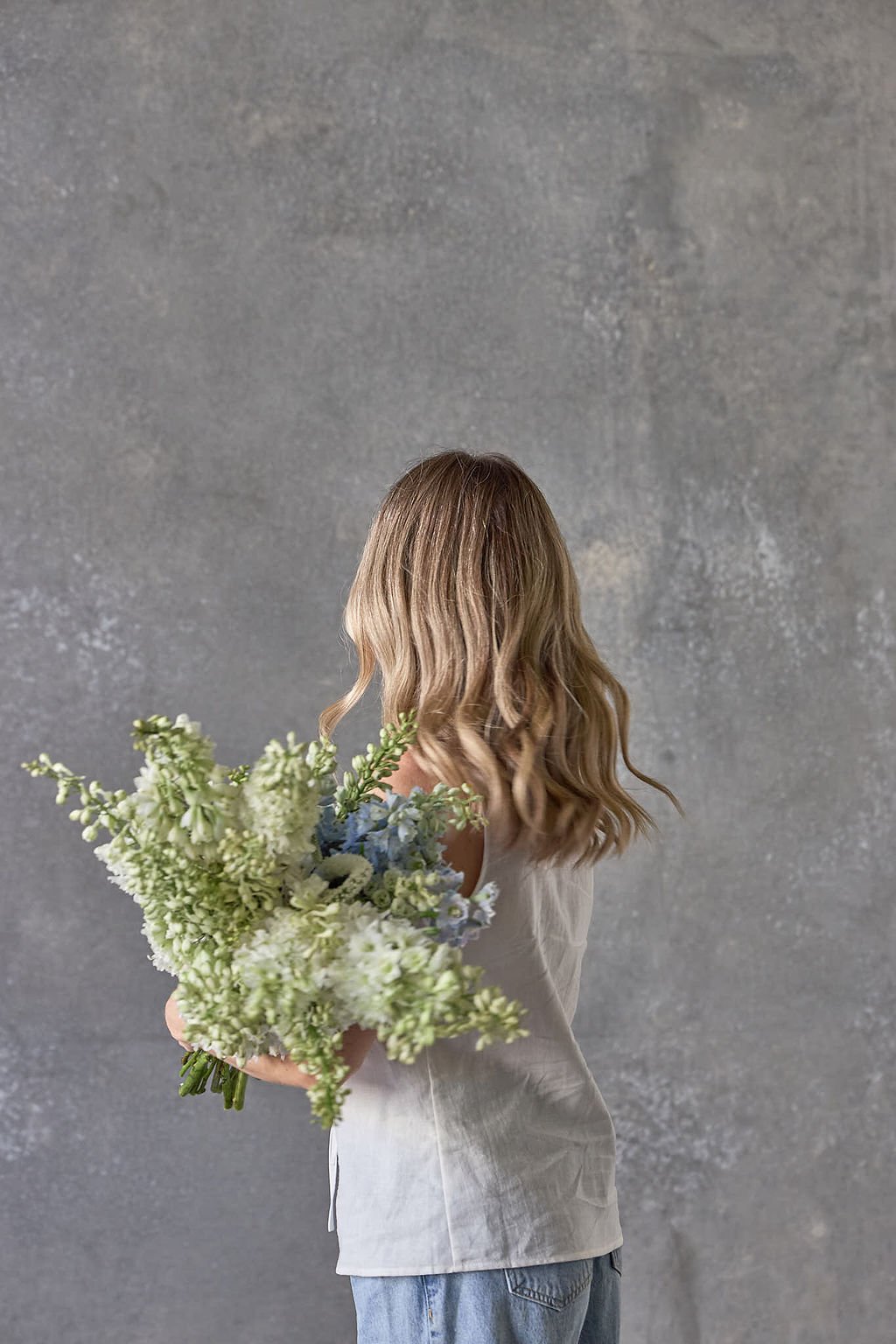 A woman with wavy blonde hair holding a bouquet of white and pale green flowers, standing against a textured gray wall, seen from the back.