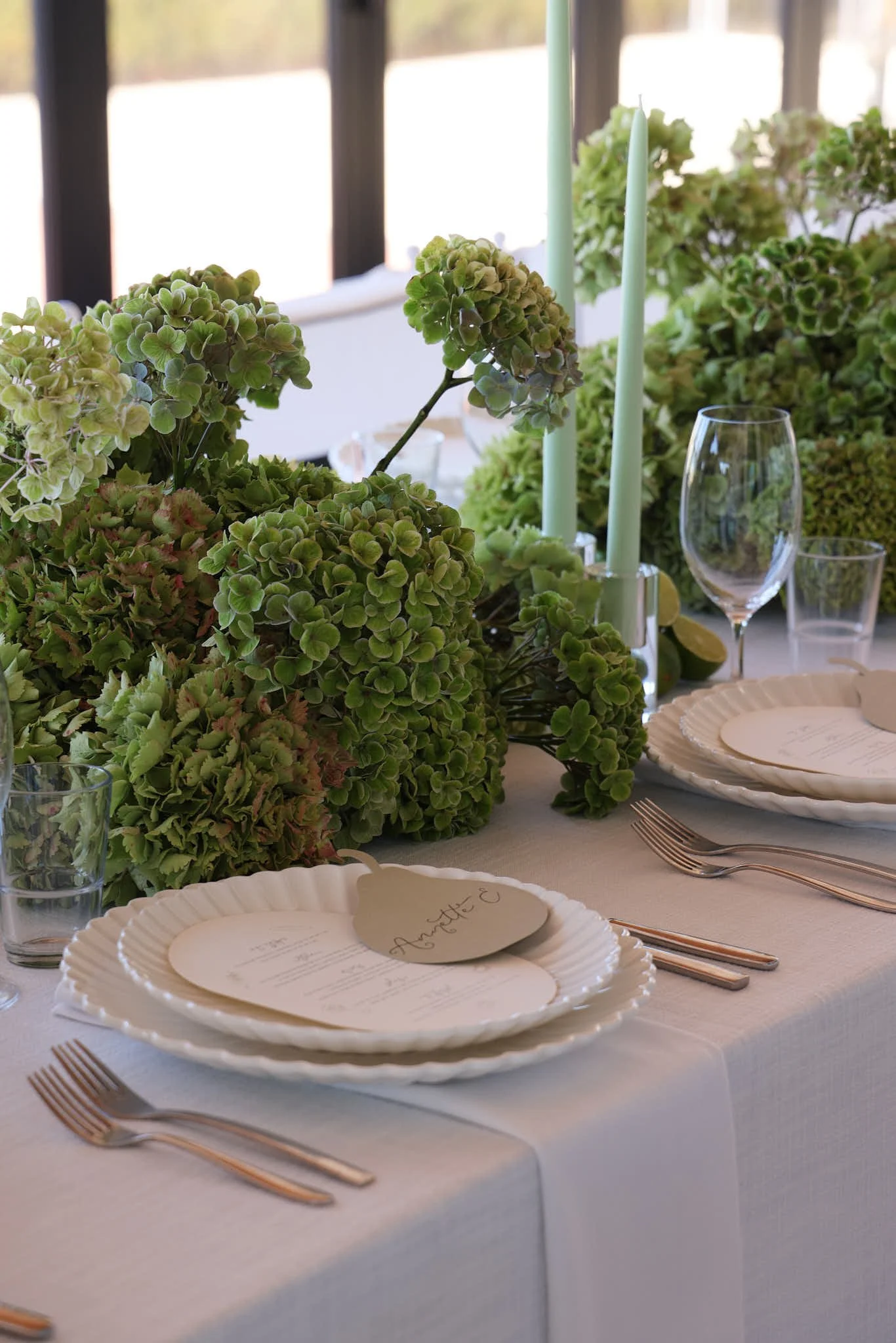 A beautifully decorated dining table with white plates, rose gold cutlery, clear glasses, and a centerpiece of green hydrangeas and candle holders, set for an event or celebration.