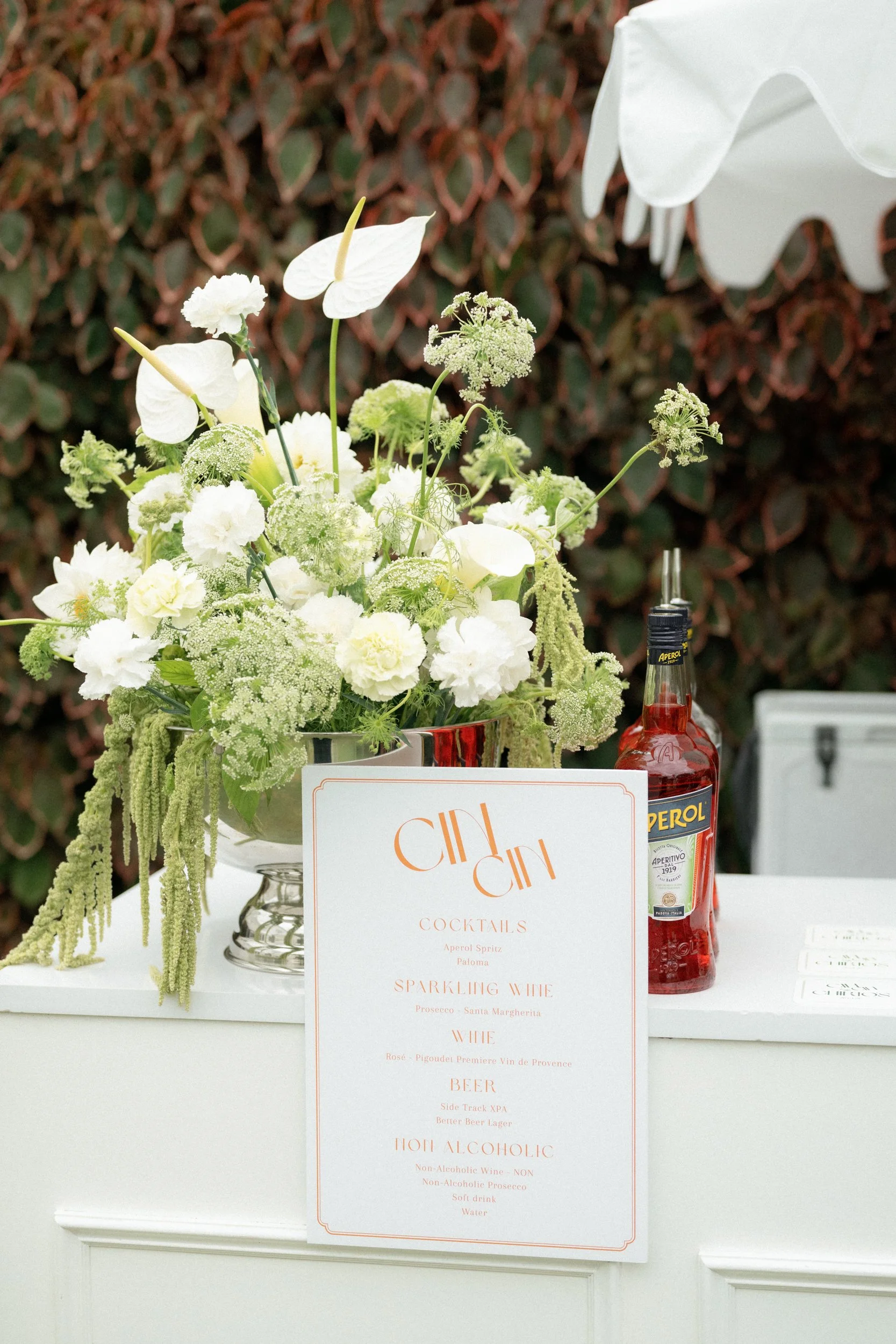 A floral arrangement with white flowers and greenery on a table, accompanied by a menu and a bottle of Aperol spritz, with a background of a hedge.
