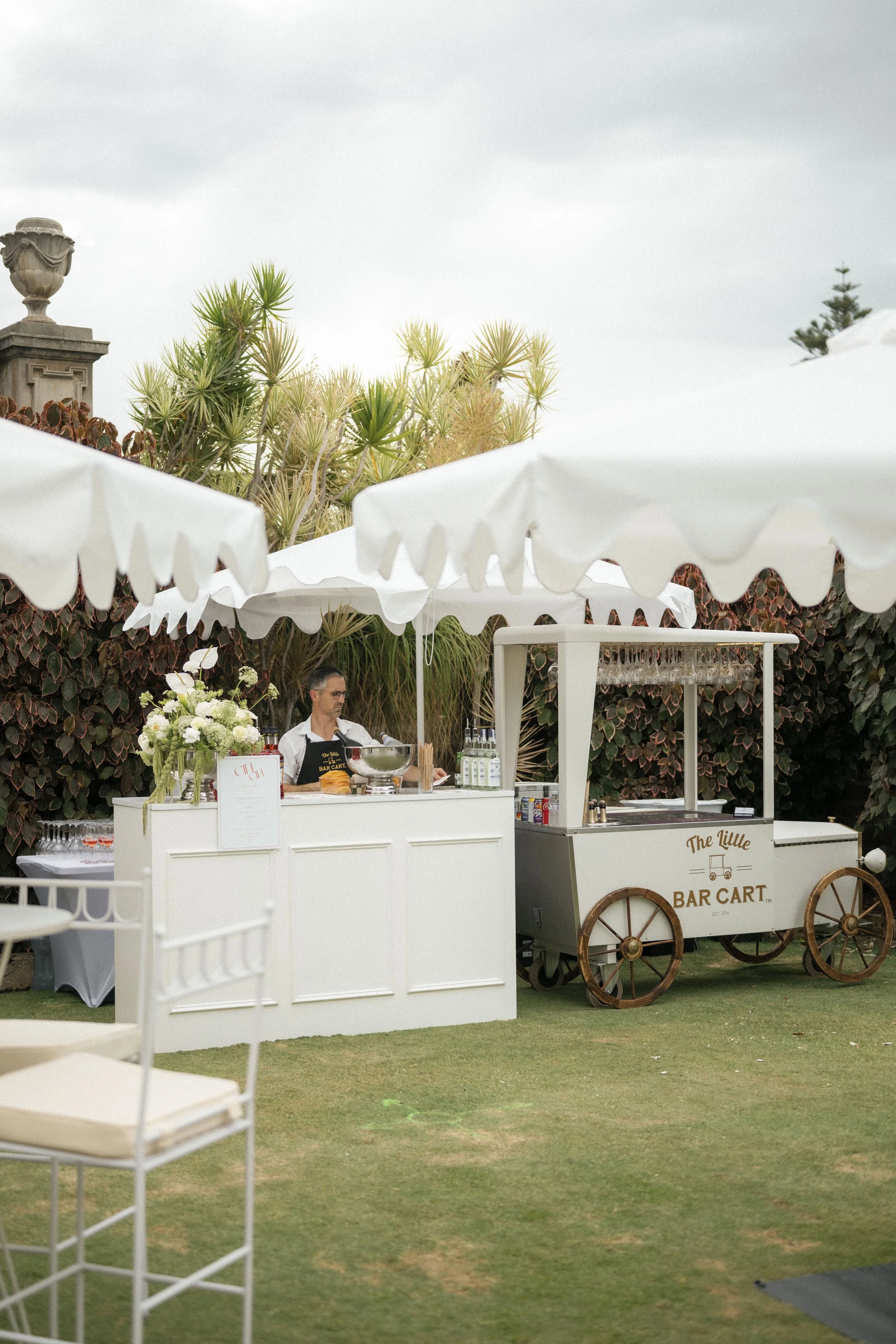 Outdoor event setup with a white bar cart labeled 'The Little Bar Cart,' a floral arrangement, umbrellas, and a bartender in a black apron under a cloudy sky, surrounded by greenery and white chairs.