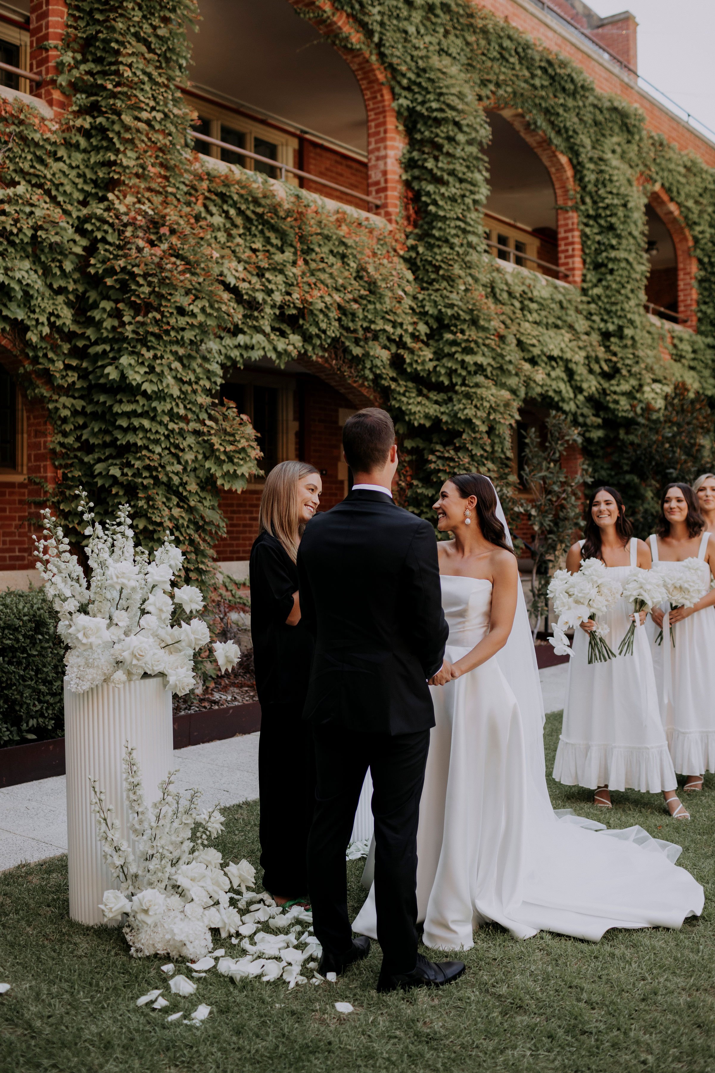 A wedding ceremony taking place outdoors with the bride and groom exchanging vows in front of a brick building covered with green vines. Bridesmaids are standing nearby holding white floral bouquets.