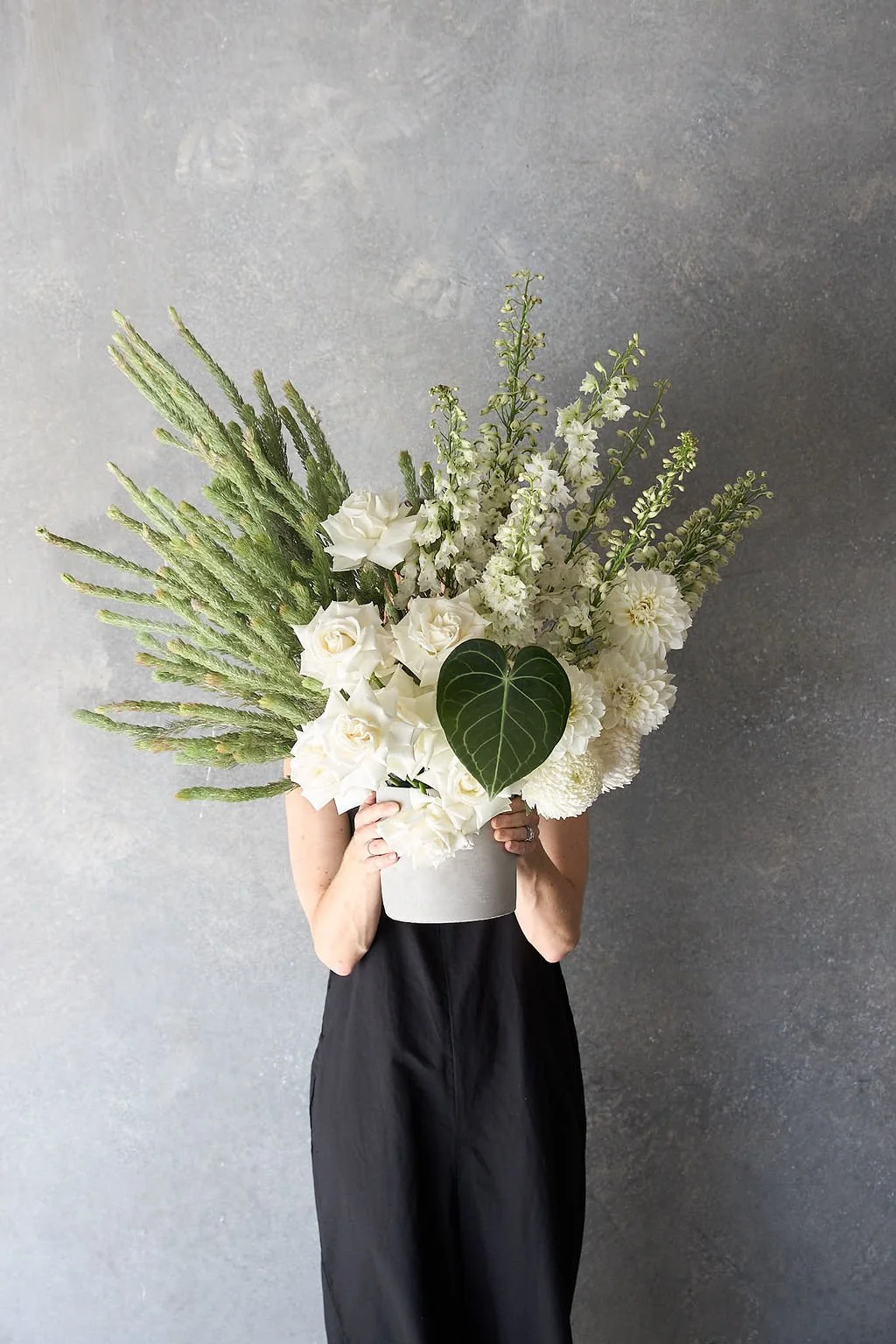 Person holding a large white floral bouquet with various white flowers and greenery in front of a gray wall.