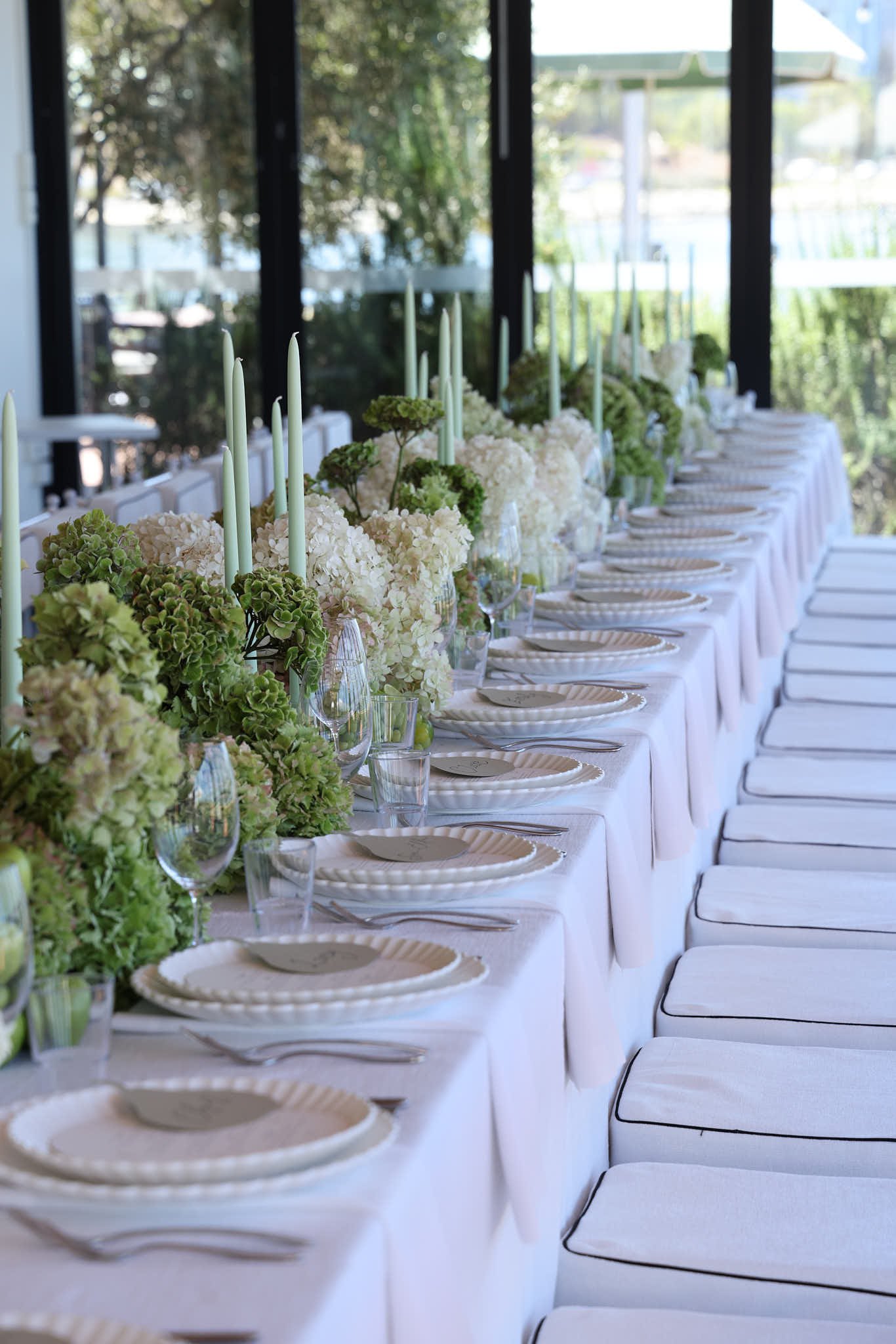 A long dining table decorated with white and green floral arrangements, white candles, and place settings in a bright room with large windows and outdoor greenery visible in the background.