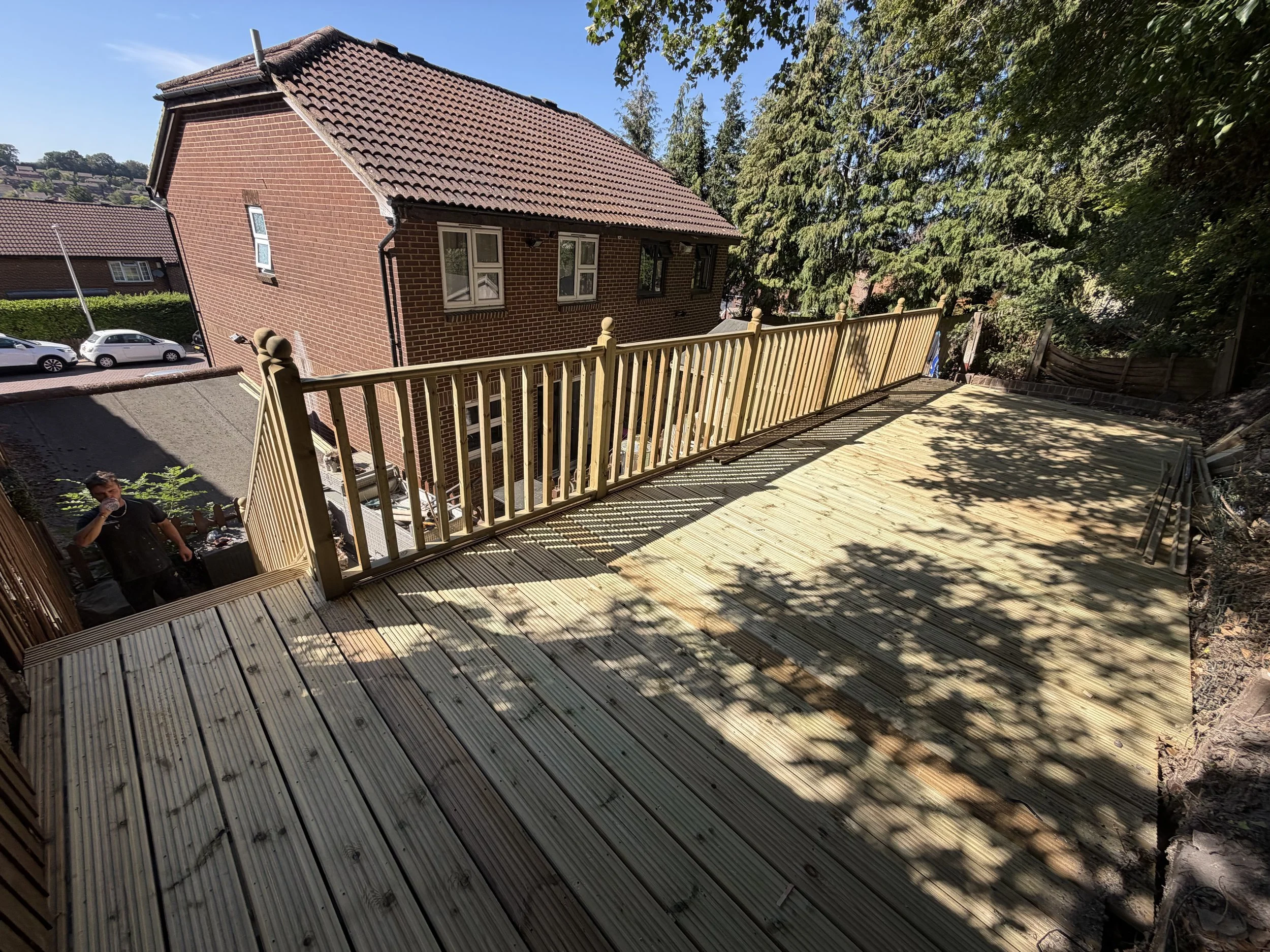 View from a wooden deck with a railing, overlooking a red brick house with a brown tiled roof, cars parked nearby, and tall trees surrounding the area on a sunny day.