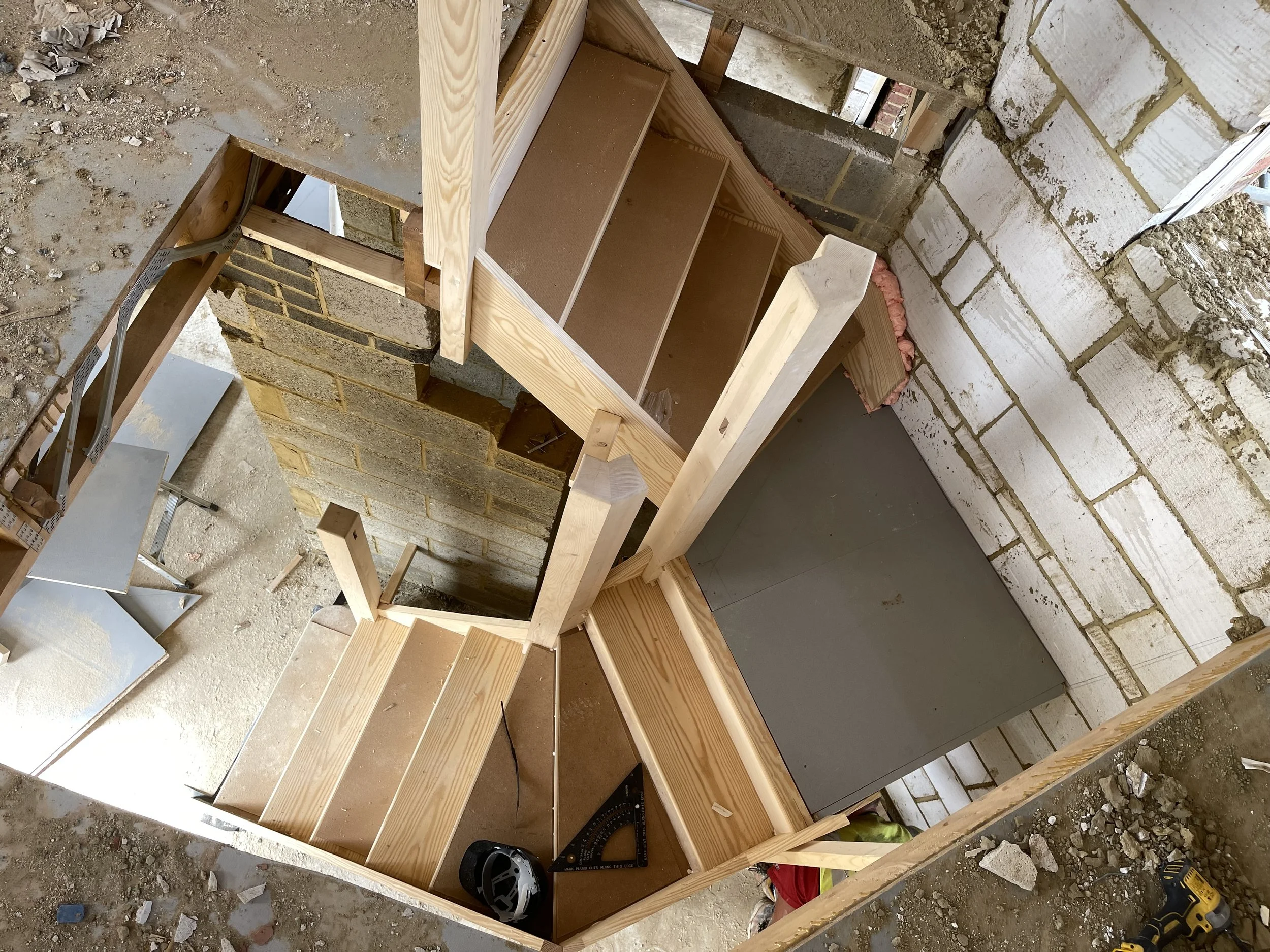 Interior view of a construction site showing wooden framing for a staircase. The structure is built on a concrete and brick wall, with construction tools and debris scattered on the floor.