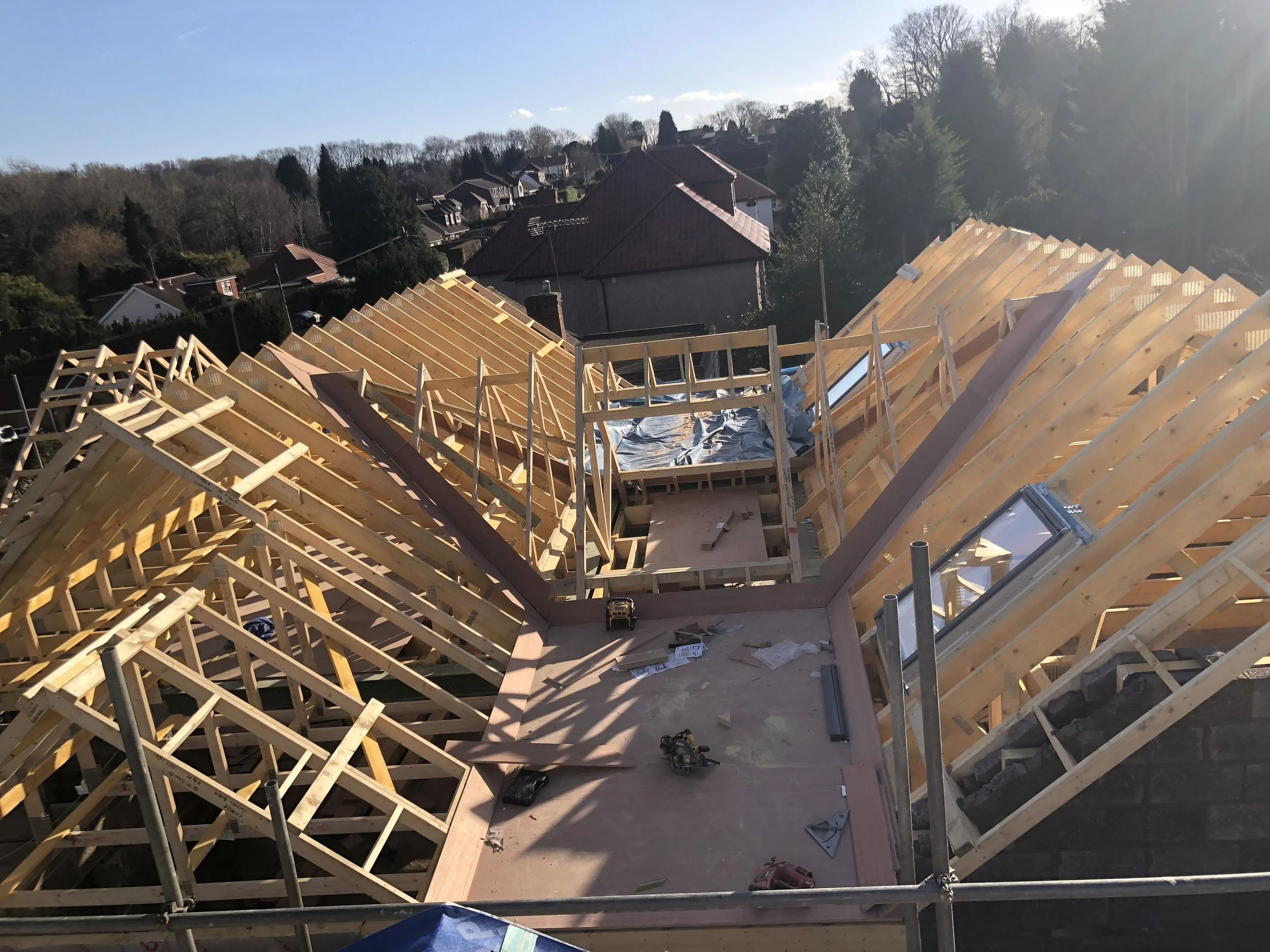 Construction site with wooden roof framing in progress on a residential building, with rooftops and trees in the background on a clear day.