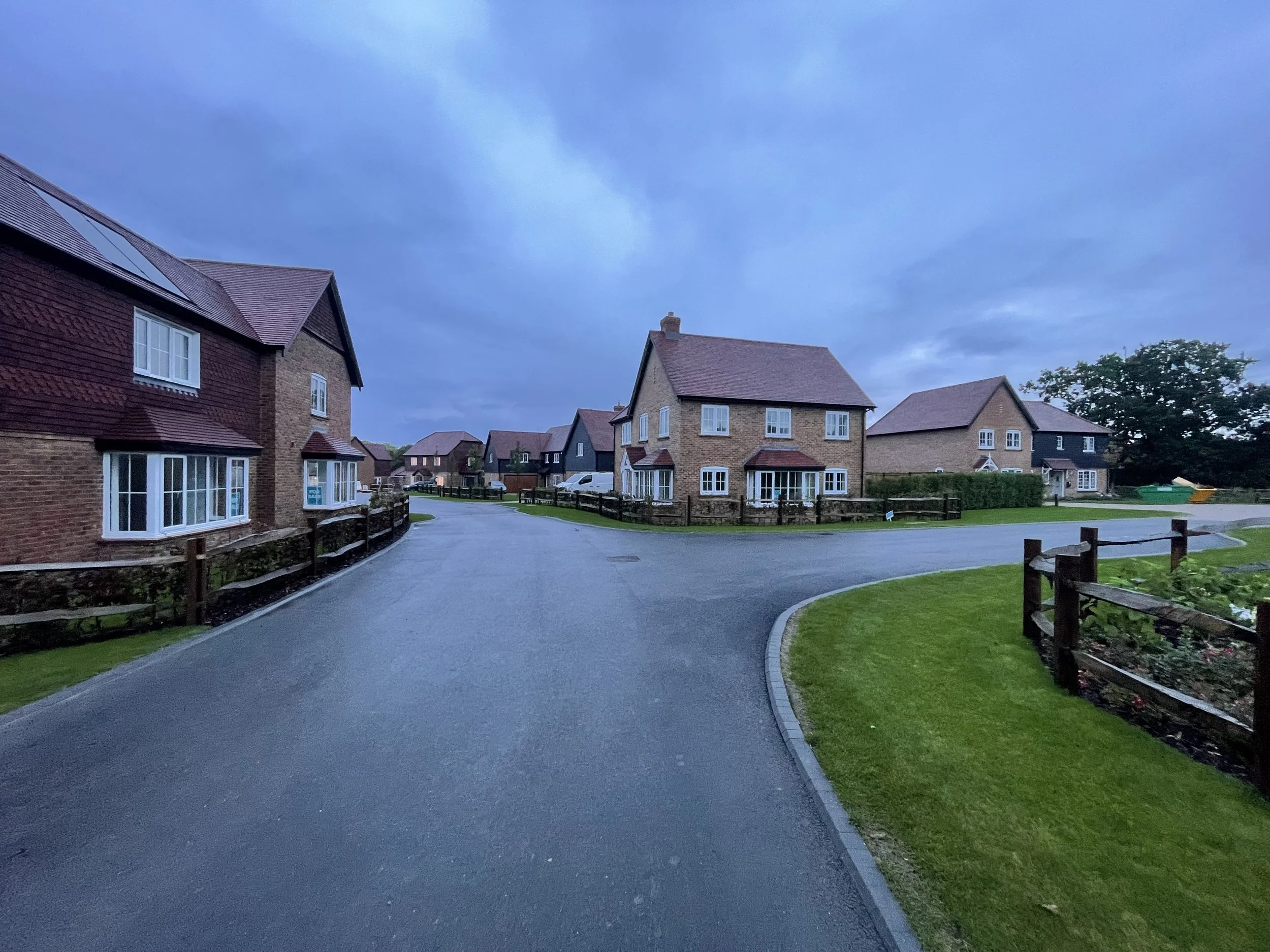 A residential neighborhood with brick and wood houses, paved roads, green lawns, and trees, on a cloudy evening.