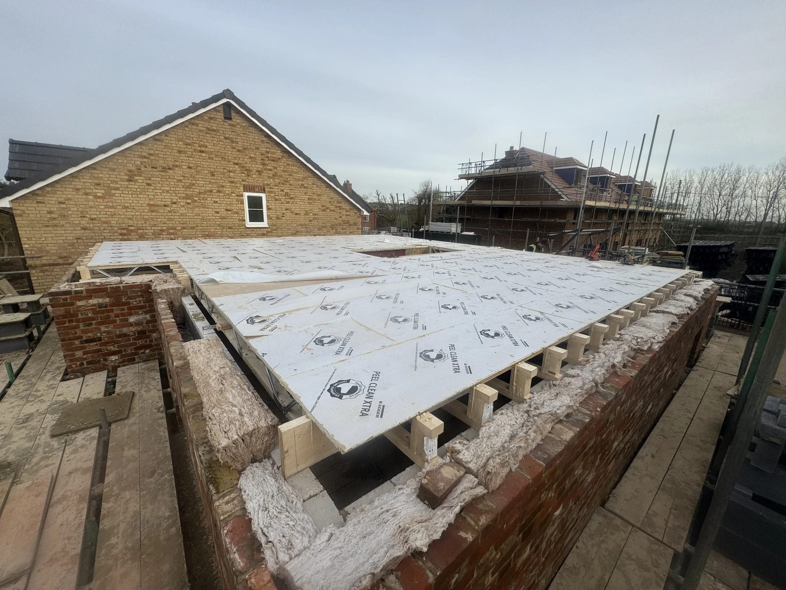 Construction site with building framework, insulation material, and scaffolding on neighboring houses with brick walls.