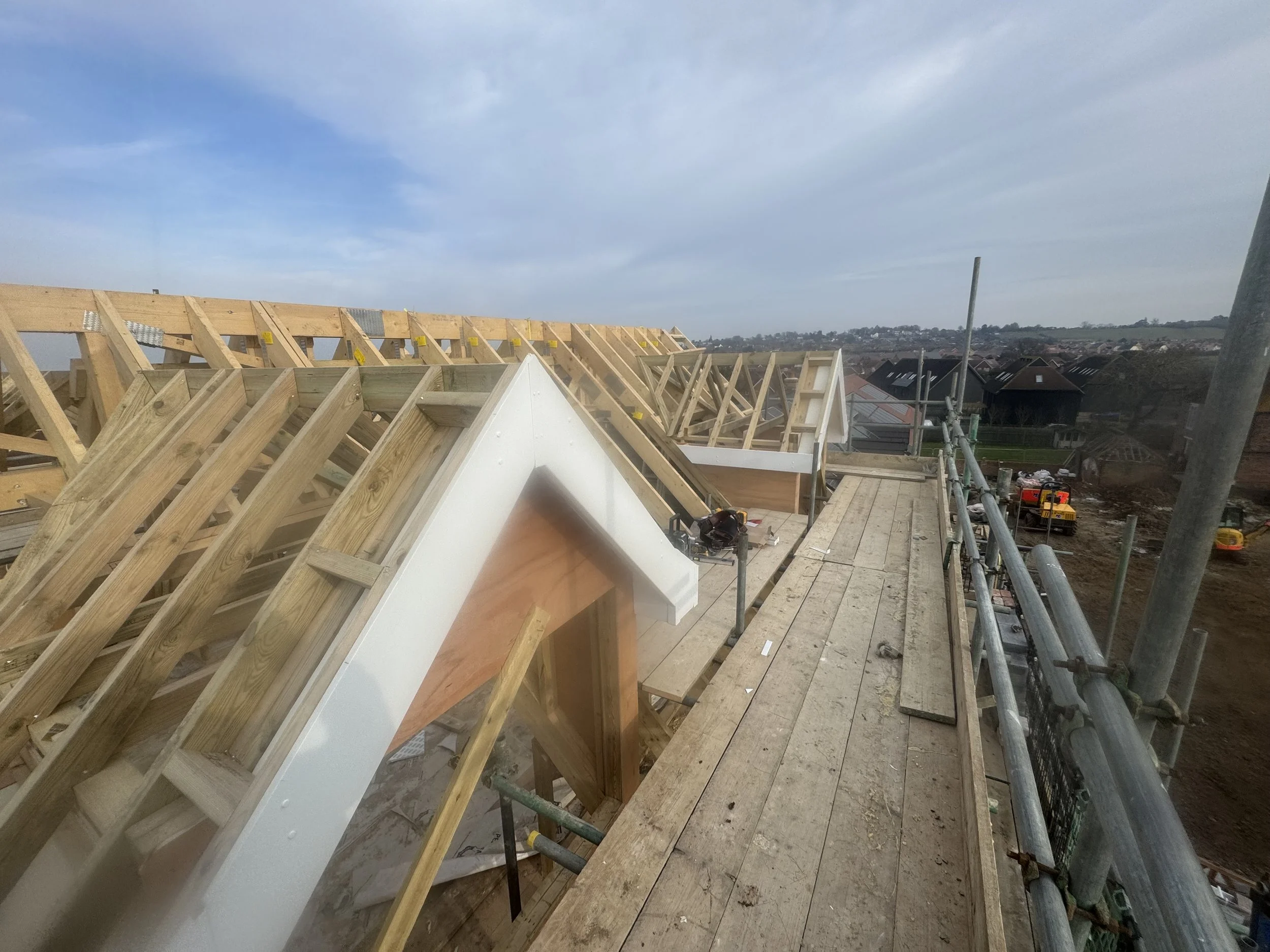 Construction site with wooden framework and scaffolding, part of a building under construction, with rooftops and cloudy sky in the background.