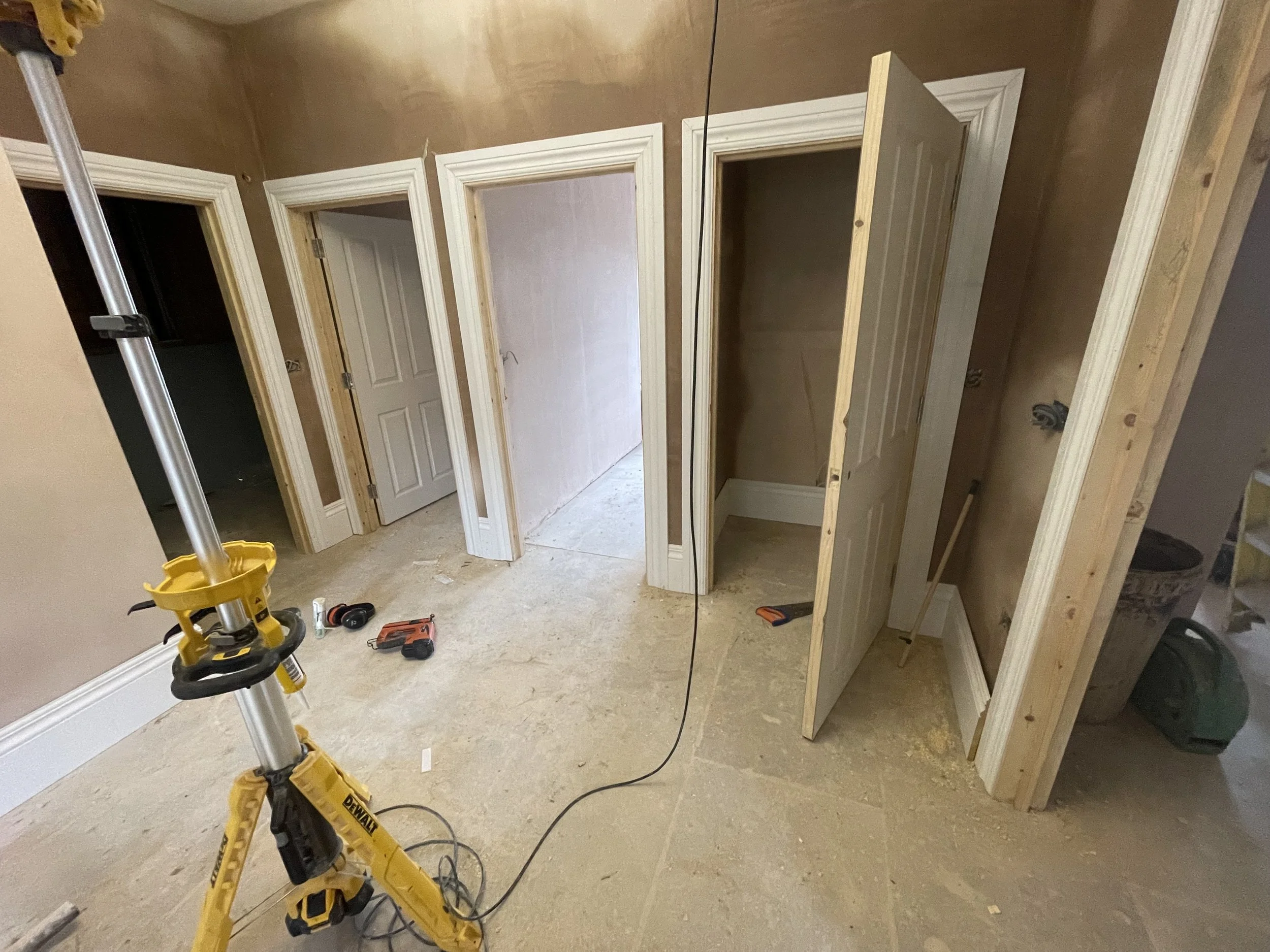 Interior of a room under construction with a floor, walls, and partially installed door frames. A yellow power tool and a drill are on the floor, and a mirror reflects part of the room.