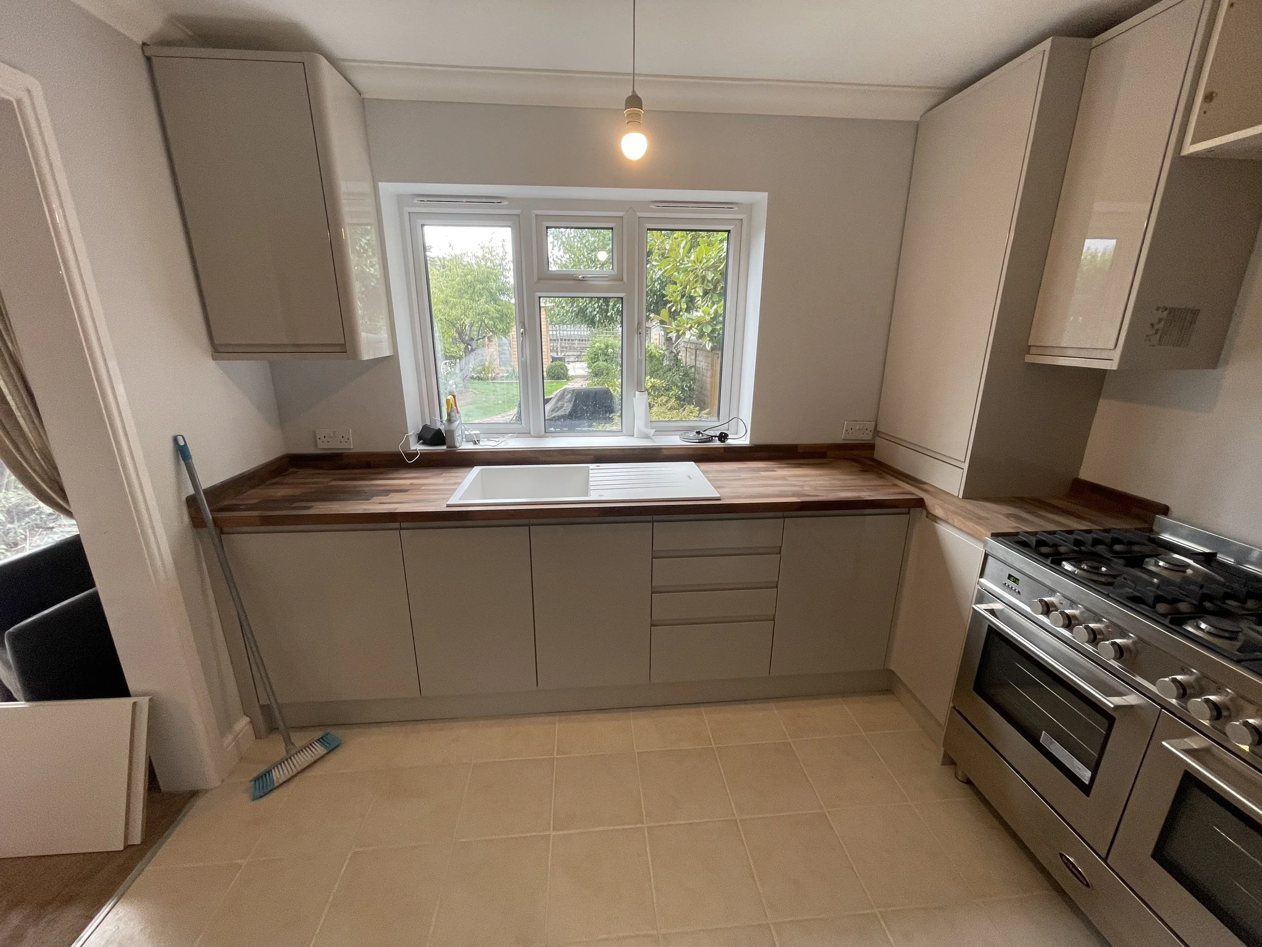 A modern kitchen with beige tile flooring and white cabinetry. There is a window in the center of the back wall showing a garden with trees and bushes. A wooden countertop with a sink is below the window. To the right, there is a stainless steel gas stove and oven. Cleaning tools, including a broom and dustpan, are leaning against the left wall.