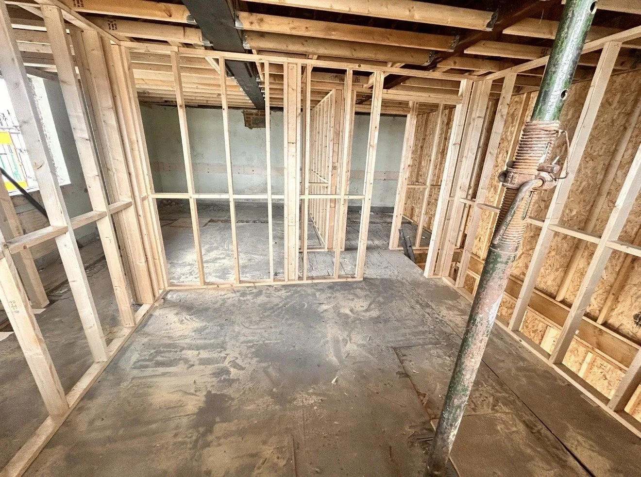 Interior view of a residential construction site with exposed wooden framing and a concrete floor, ceiling support beams, and a construction pole.