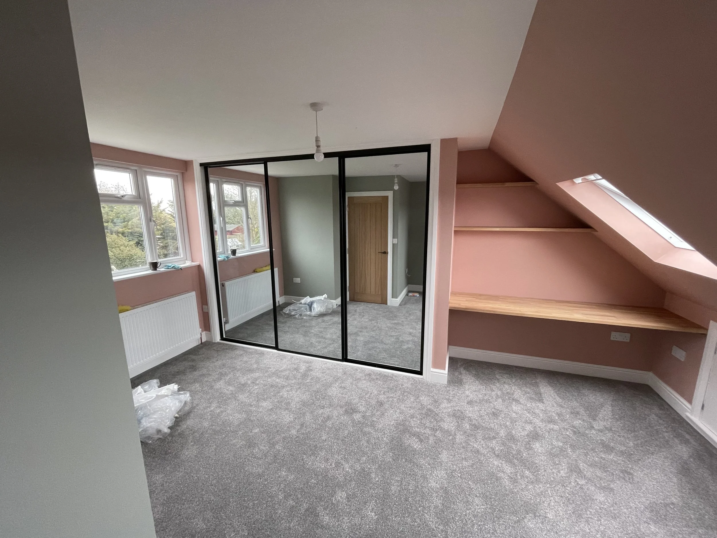 An attic room with pink walls, gray carpeting, two windows, a wooden door, and a mirror wardrobe showing a small part of the room. There are plastic bags and some items on the floor.