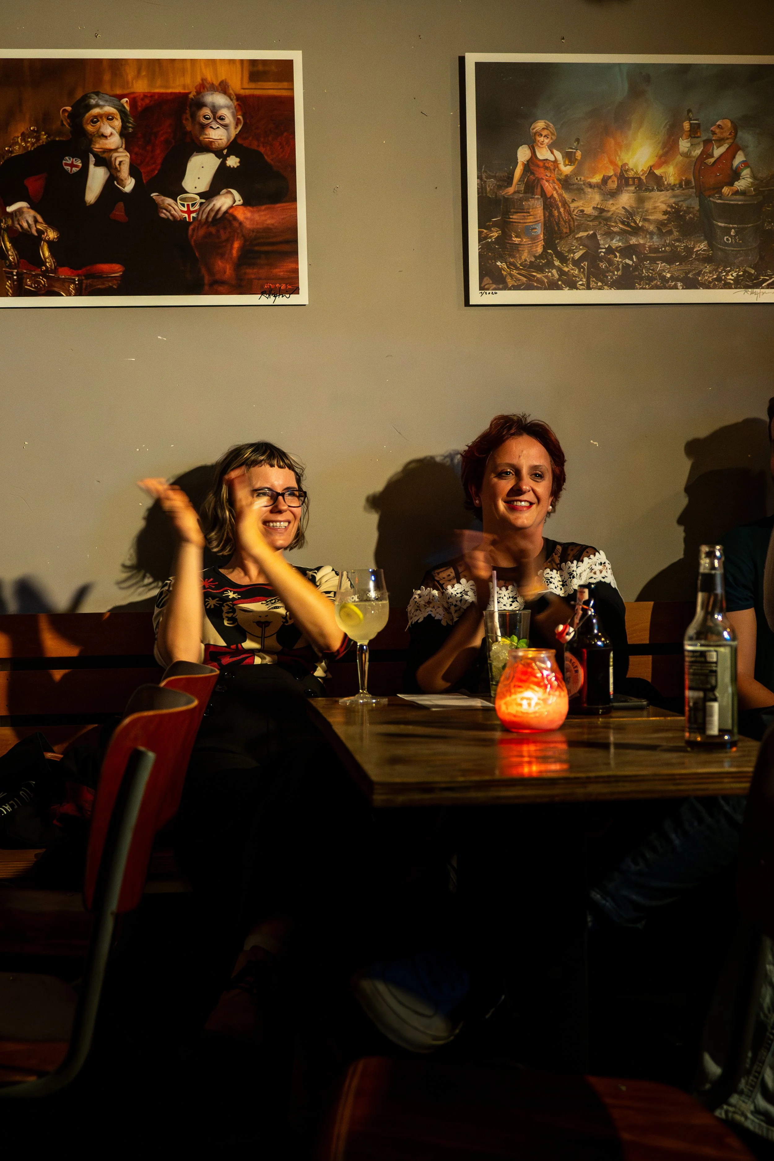 People sitting at a cocktail bar table, two women smiling and clapping, with drinks and a candle on the table. Artwork on the wall behind them.