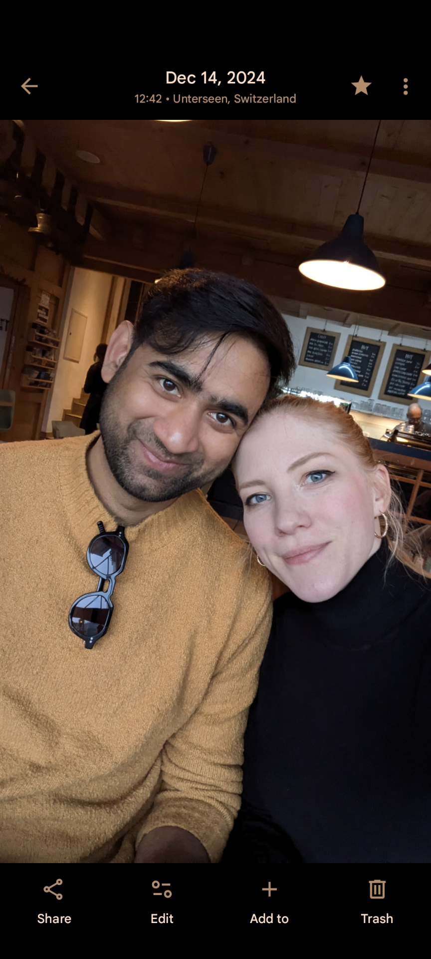 The Comedy Gallery hosts and partners Prateek, a smiling man with dark hair and wearing a mustard-colored sweater and sunglasses hanging from his collar, and Isabel, with red hair, blue eyes, and hoop earrings, posing together.